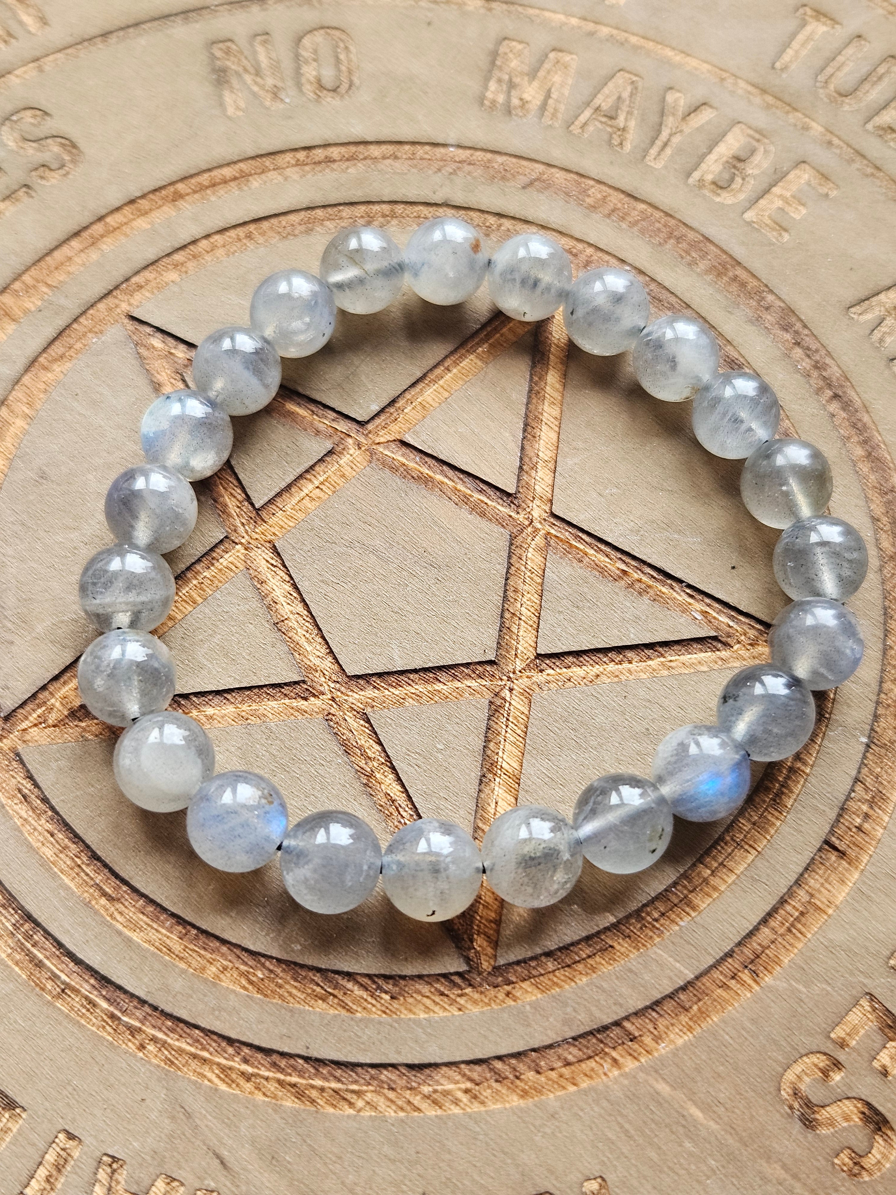 A beaded stretch bracelet made of round, translucent gray-blue gemstone beads, displayed on a wooden background featuring a carved pentagram and mystical text.