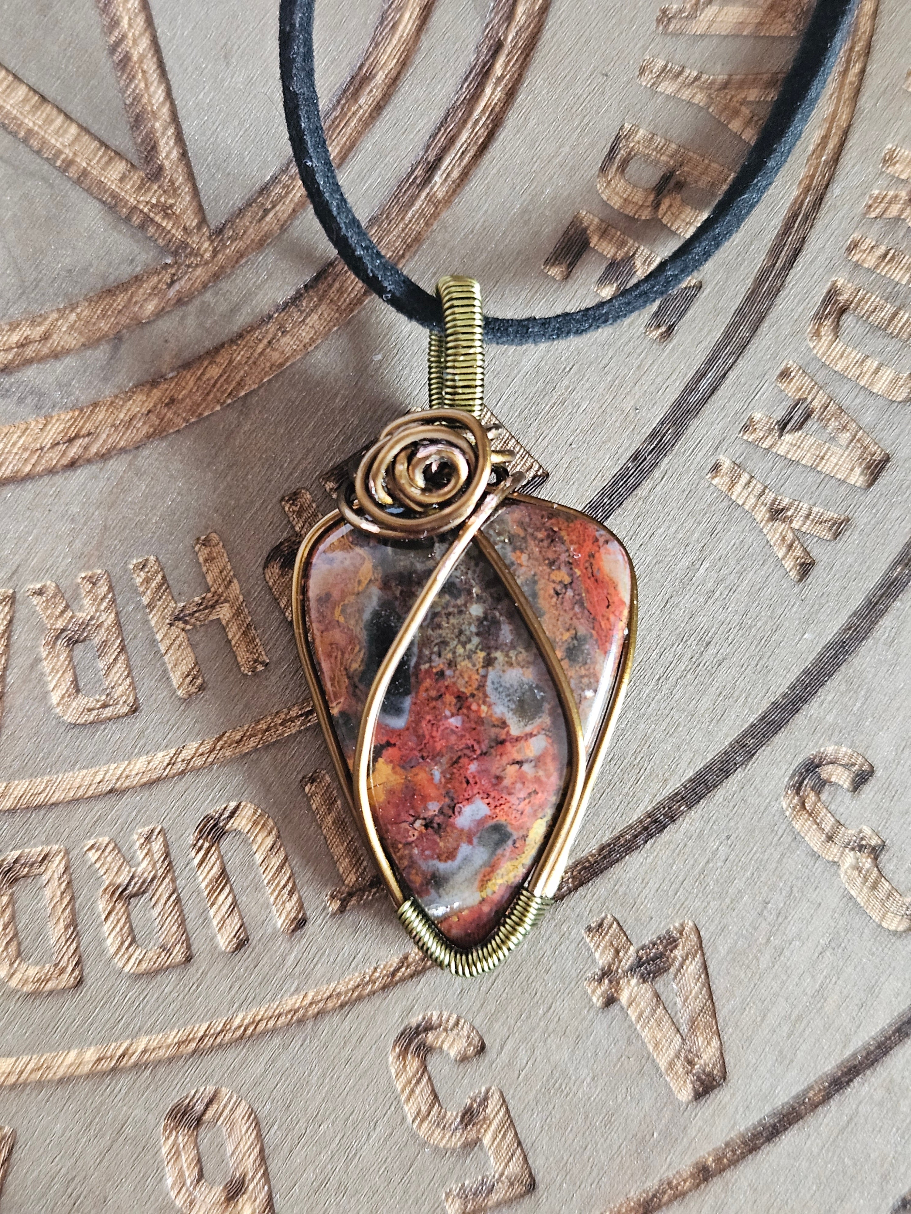 A close-up of a handmade wire-wrapped pendant necklace featuring a polished red and brown jasper stone, set against a textured wooden background with engraved symbols.