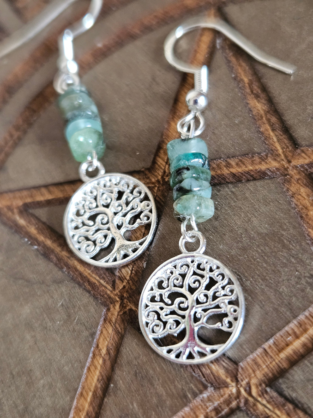 A pair of bohemian-style tree of life dangle earrings with sterling silver filigree charms and natural  Emerald beads, displayed on a dark brown wooden background.
