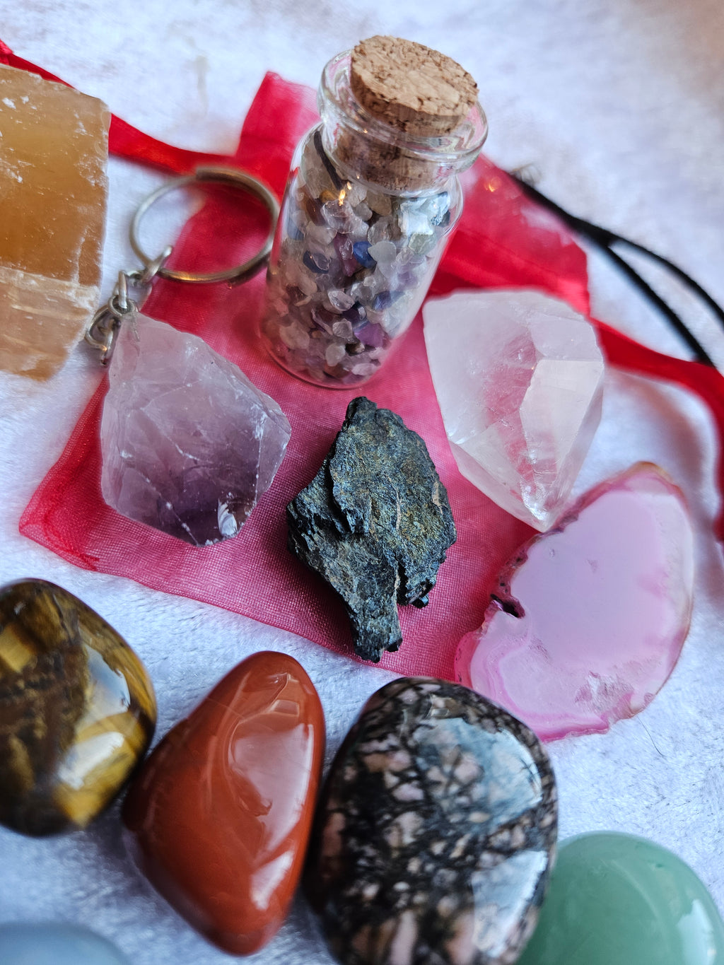 A collection of natural crystals, polished stones, and a small glass vial with crystal chips arranged on a white textured fabric background with a red organza bag.
