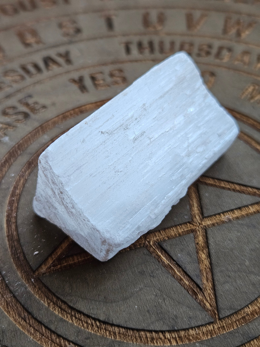 A rough, pale gray-white crystal stone with natural texture, resting on a dark wooden board engraved with a pentagram and days of the week.