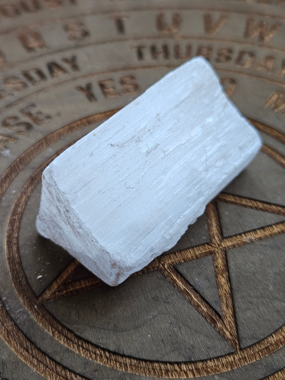 A rough, pale gray-white crystal stone with natural texture, resting on a dark wooden board engraved with a pentagram and days of the week.