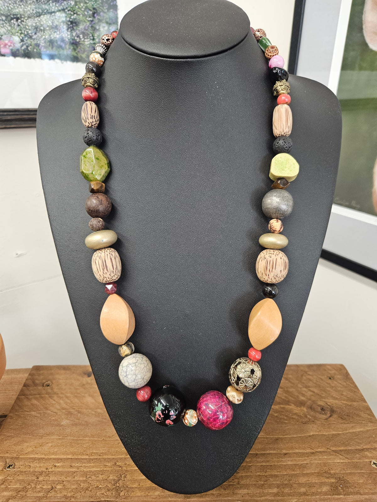 A multi-colored beaded boho necklace displayed on a black mannequin bust, featuring a variety of textured wooden beads, polished stone beads, and colorful accents on a wooden table.
