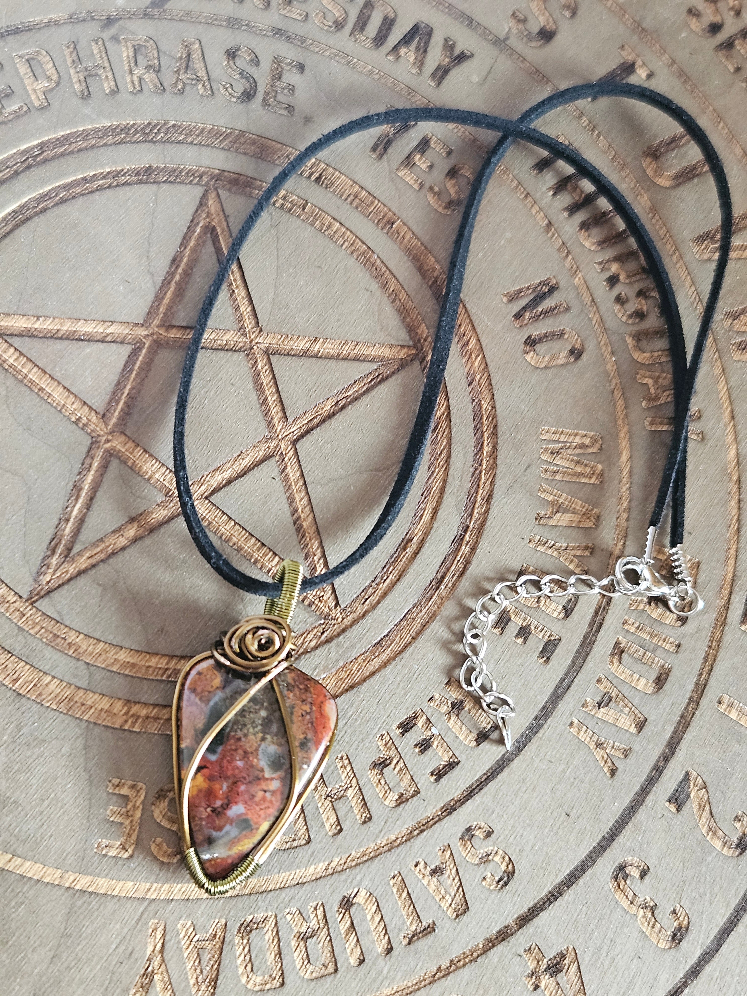 A black cord necklace with a spiral-wrapped brown and orange stone pendant, displayed on a circular wooden surface engraved with days of the week and a pentagram