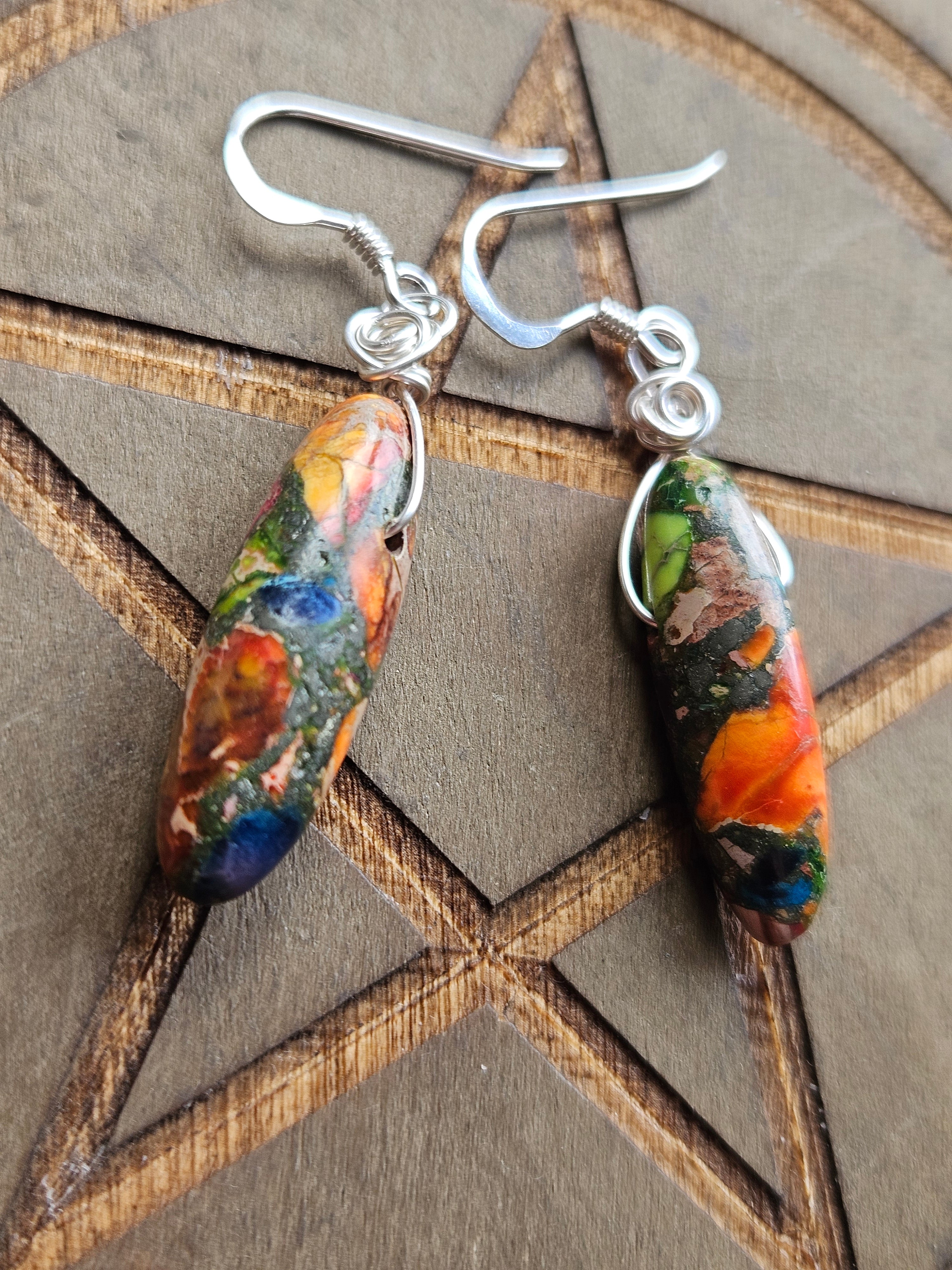 A pair of handmade dangle earrings with silver fishhook wires and teardrop-shaped Sediment Jasper beads featuring a marbled, fluid-art pattern of orange, green, blue, and brown, displayed on a wooden background with a pentagram design.