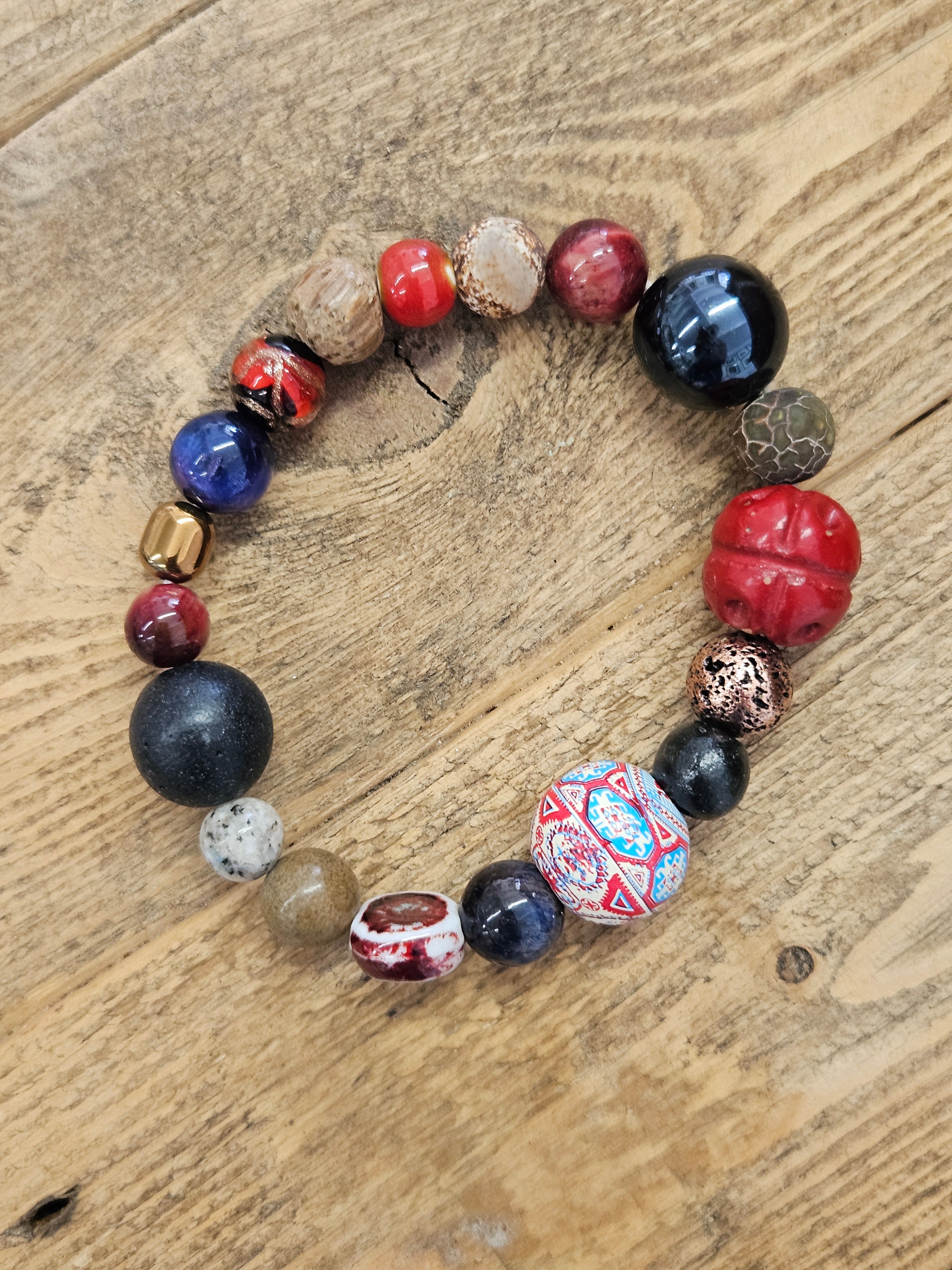 A handmade beaded bracelet made of various textured, patterned, and polished beads in red, black, blue, brown, and white, arranged in a circle on a rustic wooden surface.