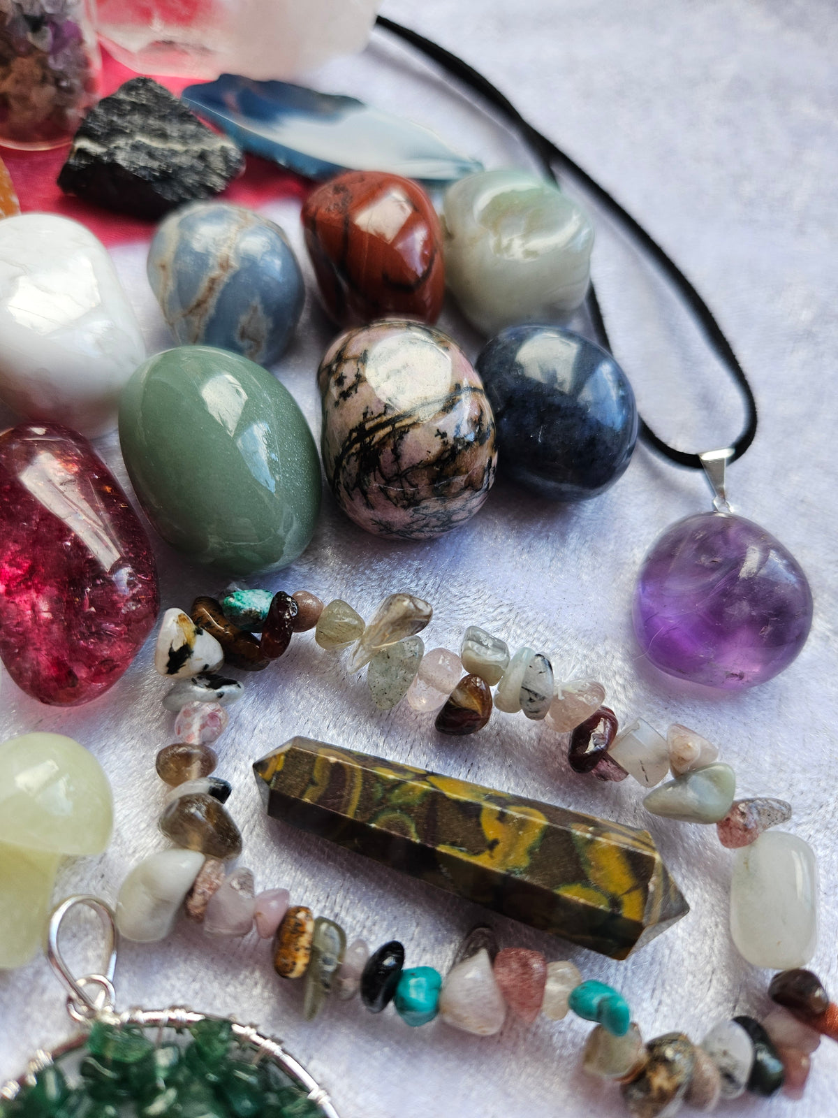 A collection of polished and tumbled crystals, gemstones, and jewelry pieces arranged on a light textured fabric background, including a black cord necklace with a purple stone pendant, a pointed yellow-brown crystal point, a beaded bracelet, and several other colorful stones.
