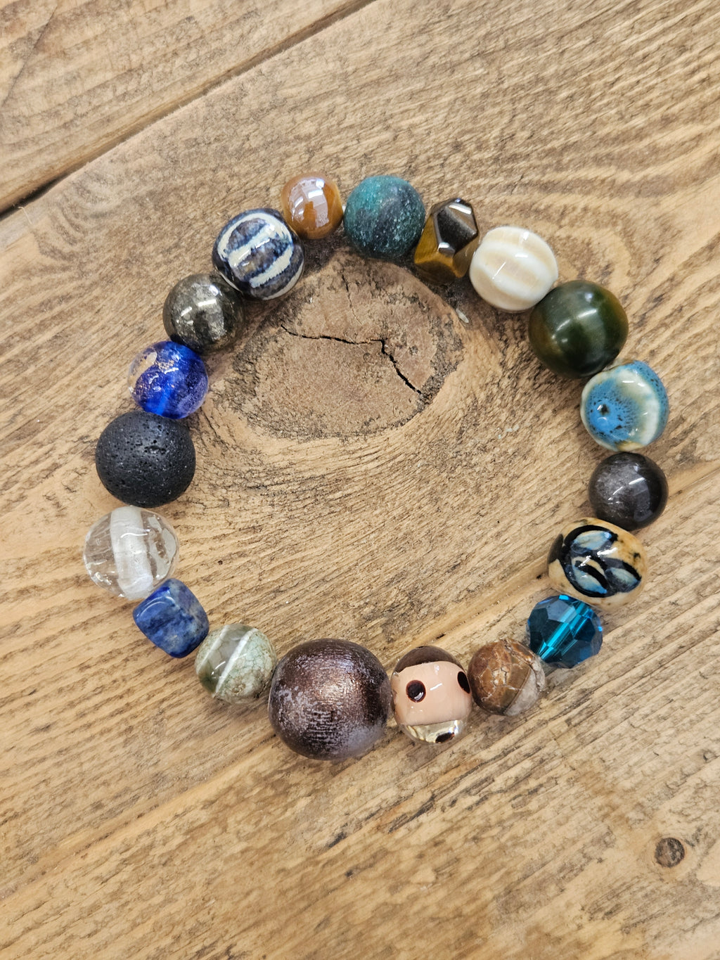 A beaded stretch bracelet made of various colorful, textured beads including blue, green, brown, black, and white, displayed on a rustic wooden surface.