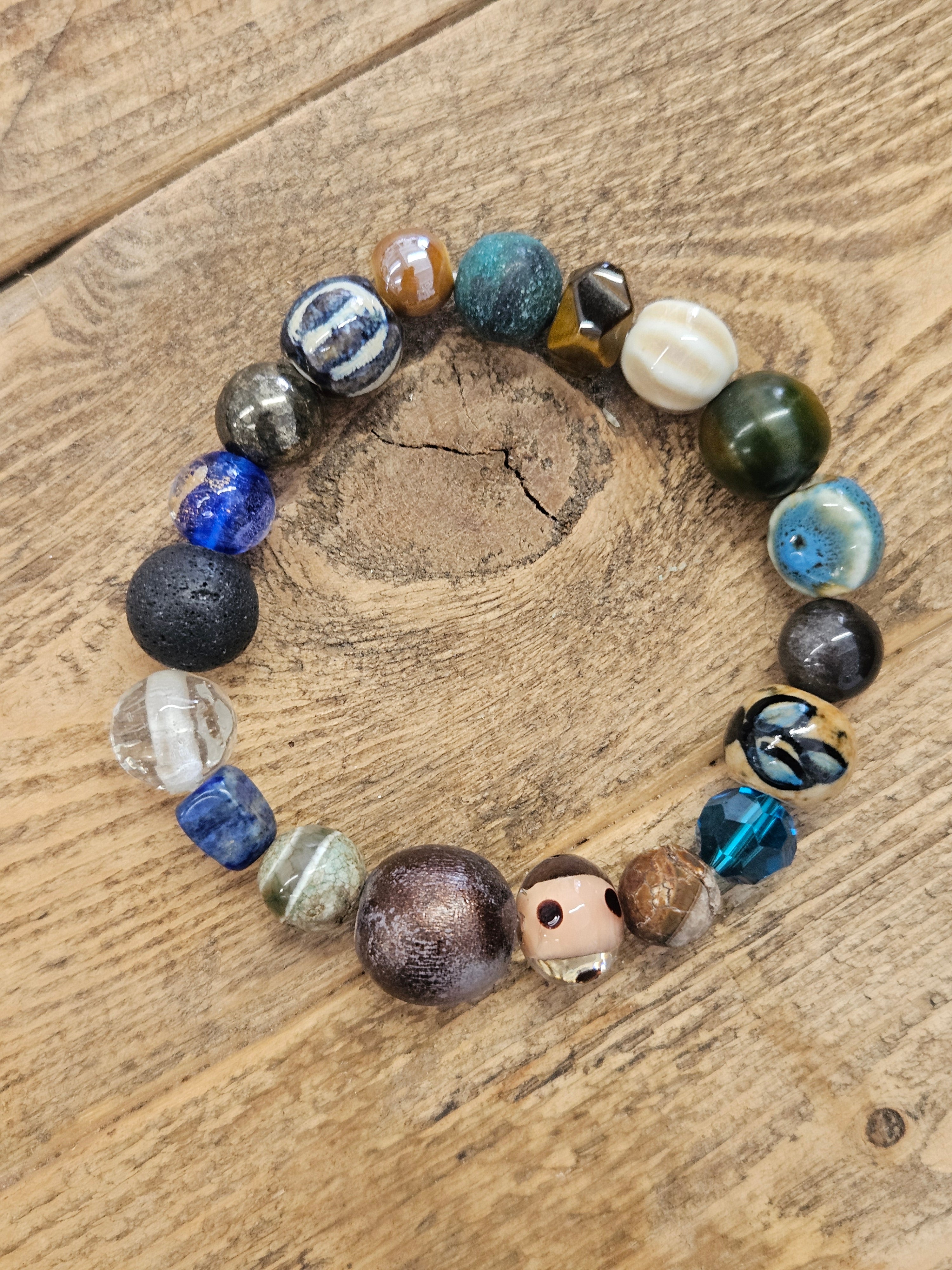 A beaded stretch bracelet made of various colorful, textured beads including blue, green, brown, black, and white, displayed on a rustic wooden surface.