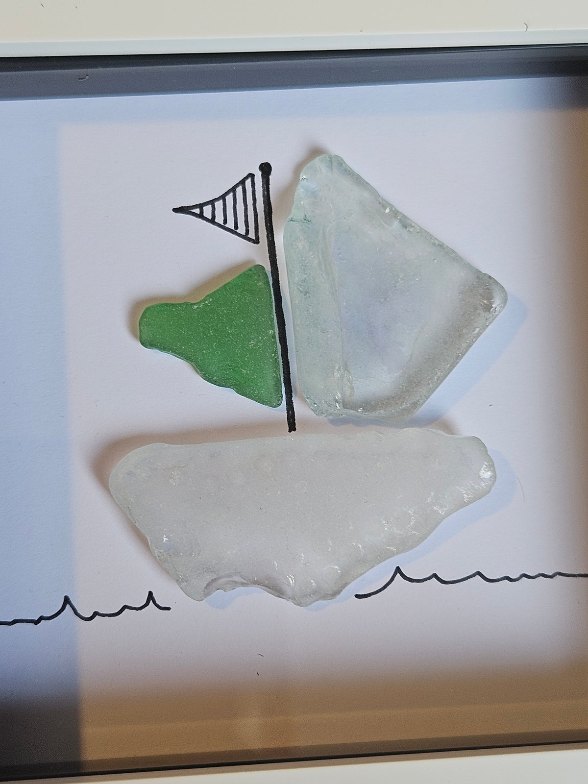A framed piece of nautical wall art featuring sea glass, with a large translucent white piece forming the hull, a smaller green piece as a sail, and another pale translucent piece as a sail, set against a white background with simple black wave lines at the bottom.