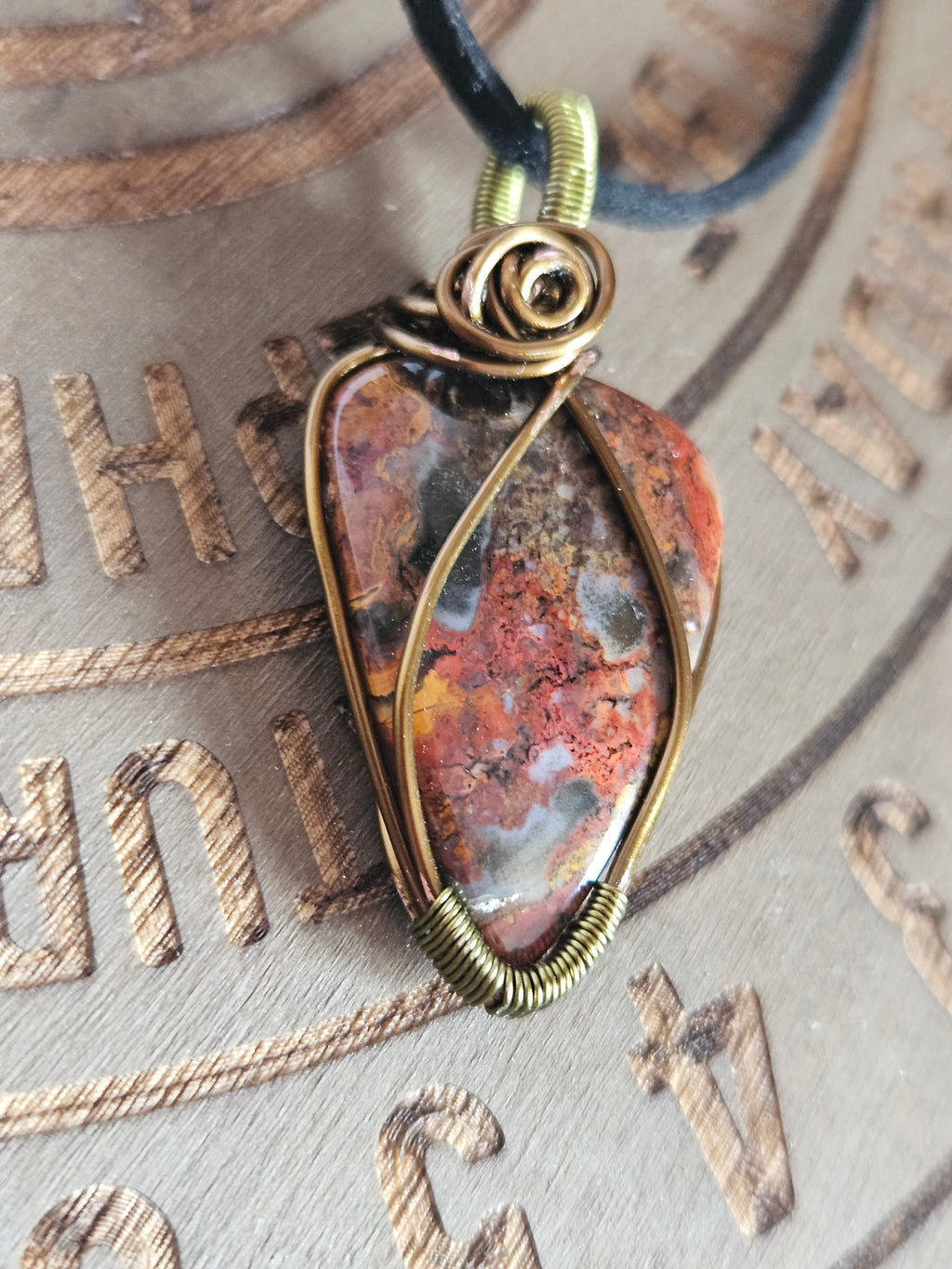 A close-up of a handmade wire-wrapped pendant necklace featuring a polished red and orange jasper stone, set against a textured, engraved brown background.