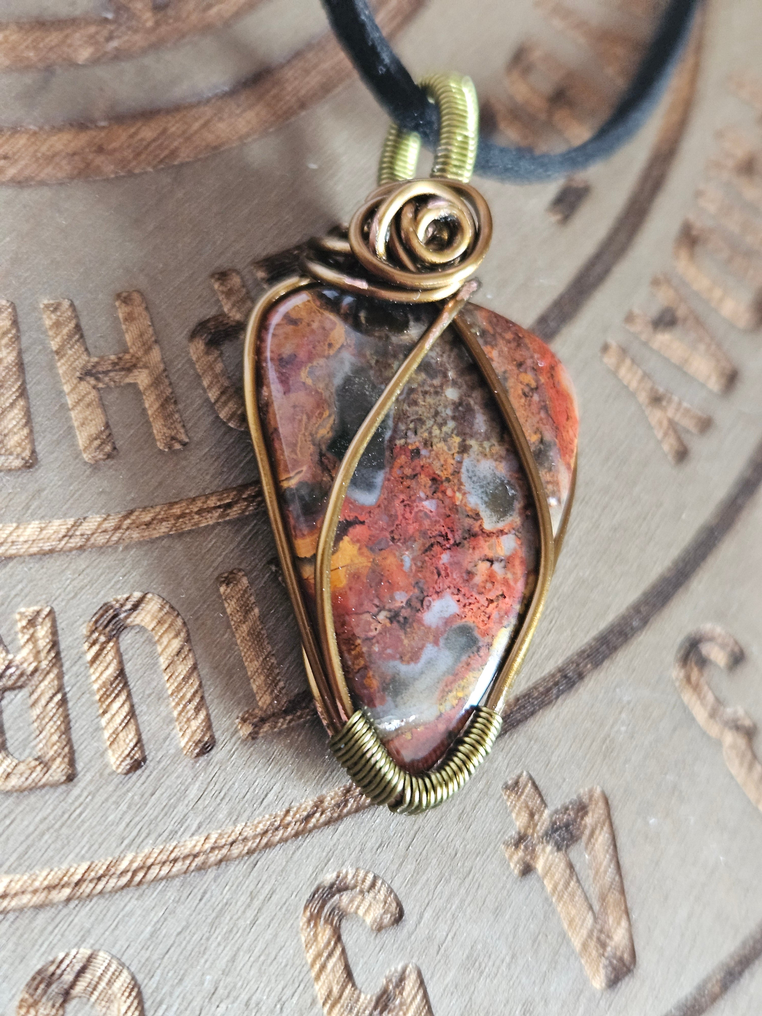 A close-up of a handmade wire-wrapped pendant necklace featuring a polished red and orange jasper stone, set against a textured, engraved brown background.