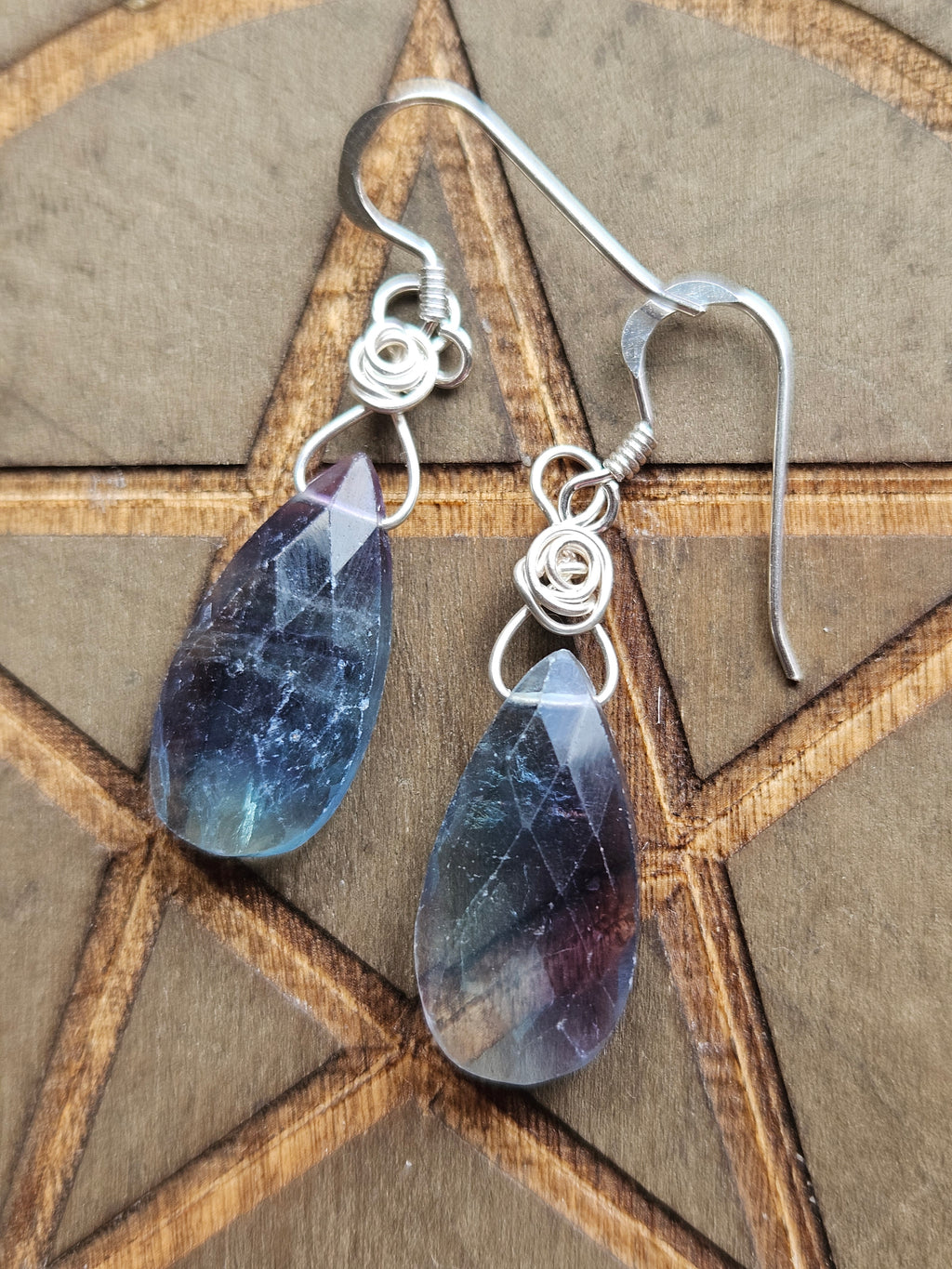 A pair of silver wire-wrapped dangle earrings with faceted teardrop-shaped blue and purple  Fluorite stones, displayed on a wooden pentagram background.