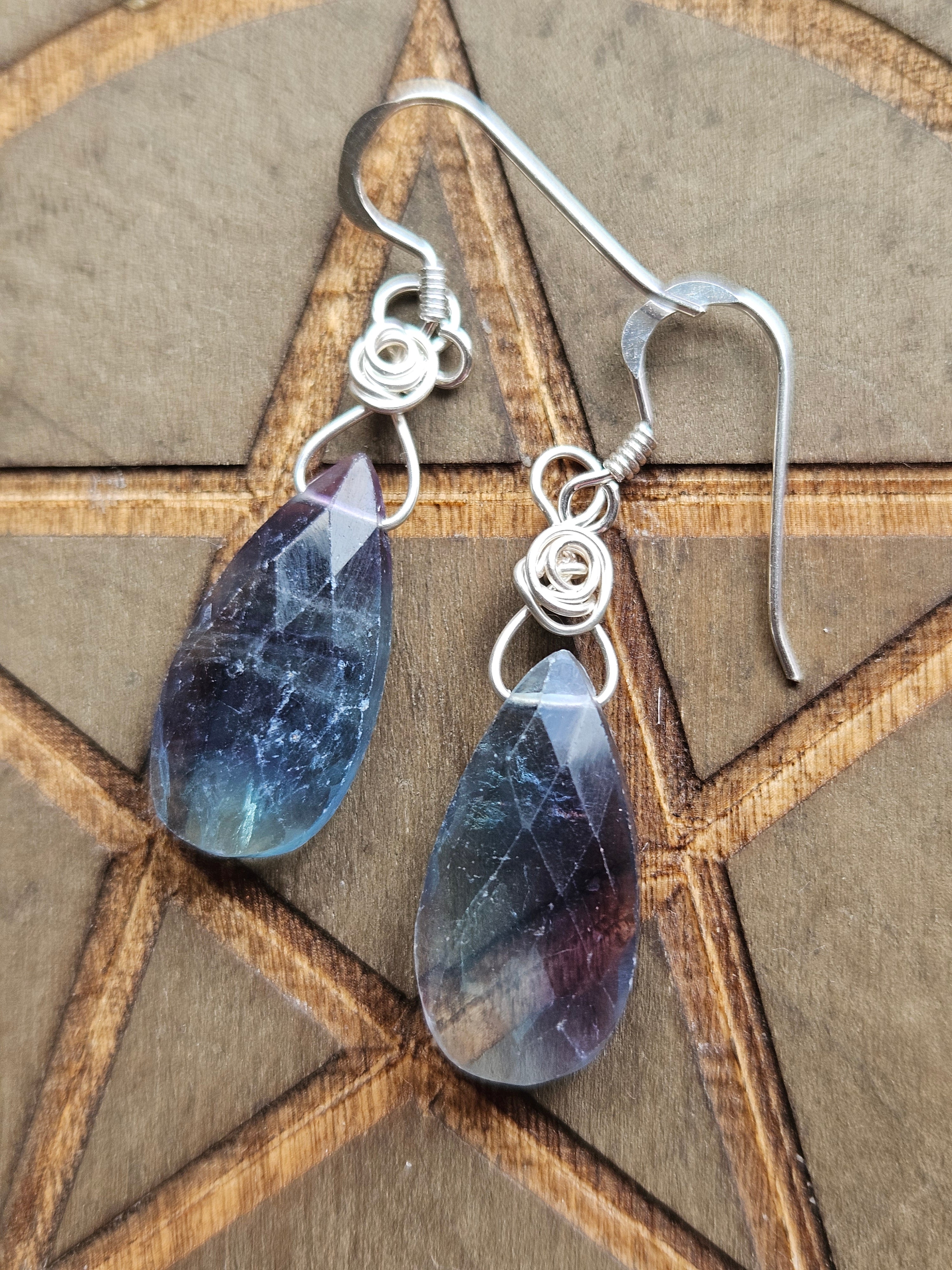 A pair of silver wire-wrapped dangle earrings with faceted teardrop-shaped blue and purple  Fluorite stones, displayed on a wooden pentagram background.