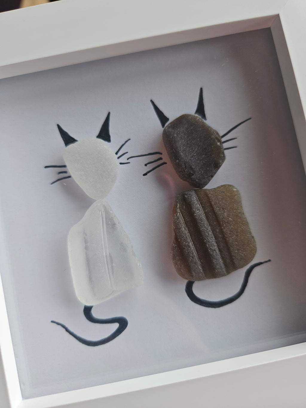 A framed piece of minimalist sea-glass art featuring two stylized cat silhouettes made from natural stones and black hand-drawn lines on a white background.