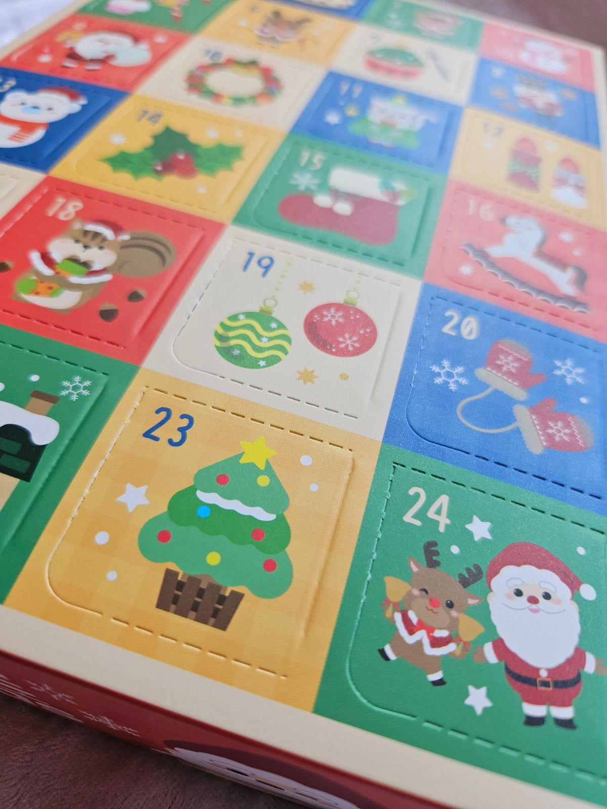 A close-up of a colorful Christmas advent calendar showing numbered square doors with festive illustrations including Santa, a Christmas tree, ornaments, and a reindeer.