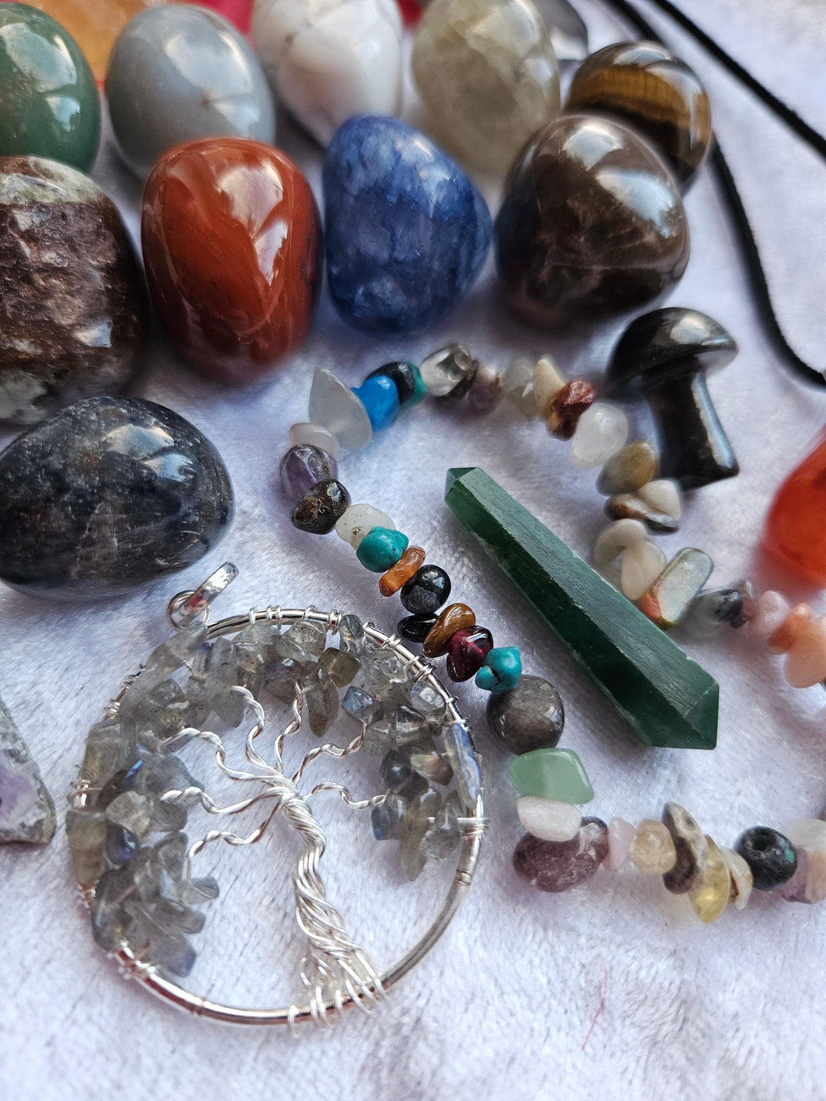 A close-up of a beaded necklace with a silver tree of life pendant, displayed alongside various polished and raw gemstones on a white textured fabric background.