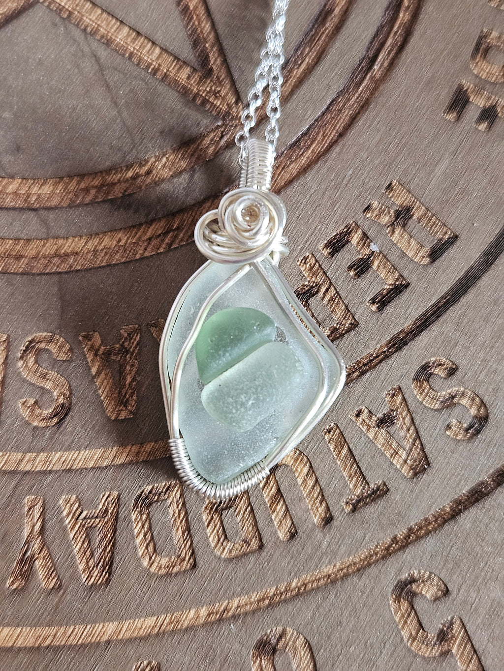 A silver-wrapped sea glass pendant necklace with a light green translucent stone, resting on a textured brown leather background with engraved lettering.
