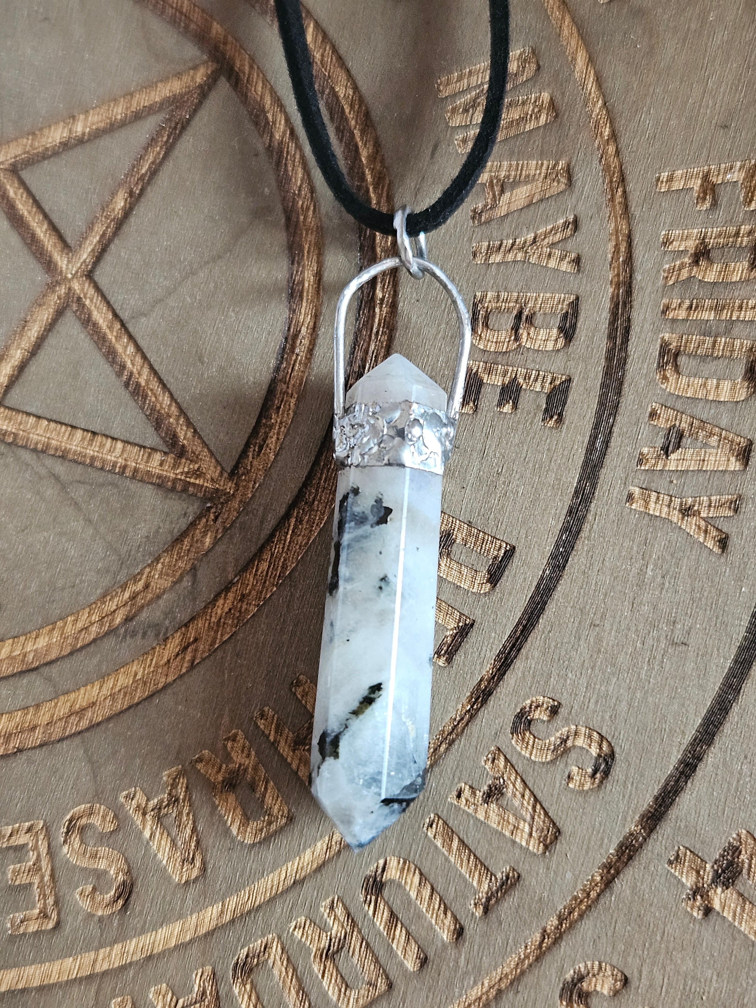 A white and black marbled crystal point pendant necklace with a silver soft soldered top and black cord, resting on a wooden engraved surface.