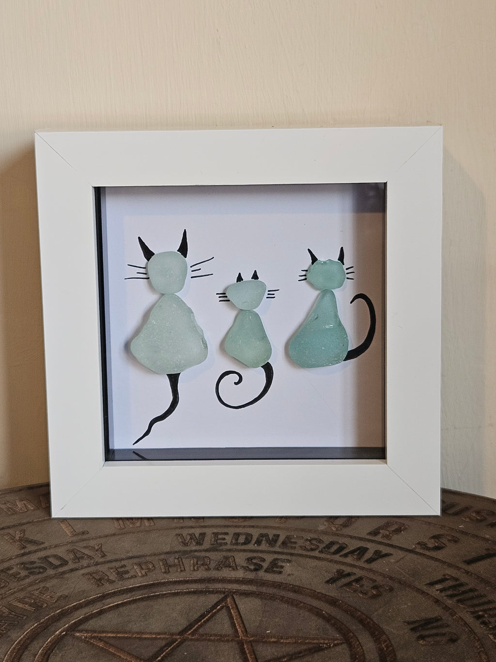A framed piece of wall art featuring three stylized sea glass cat silhouettes in pale mint green and black, displayed in a white square frame on a tabletop against a beige wall.