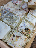 Several square handmade wax melts in off-white and cream tones, embedded with dried botanicals, seeds, and herbs, arranged in shredded paper inside a cardboard box.