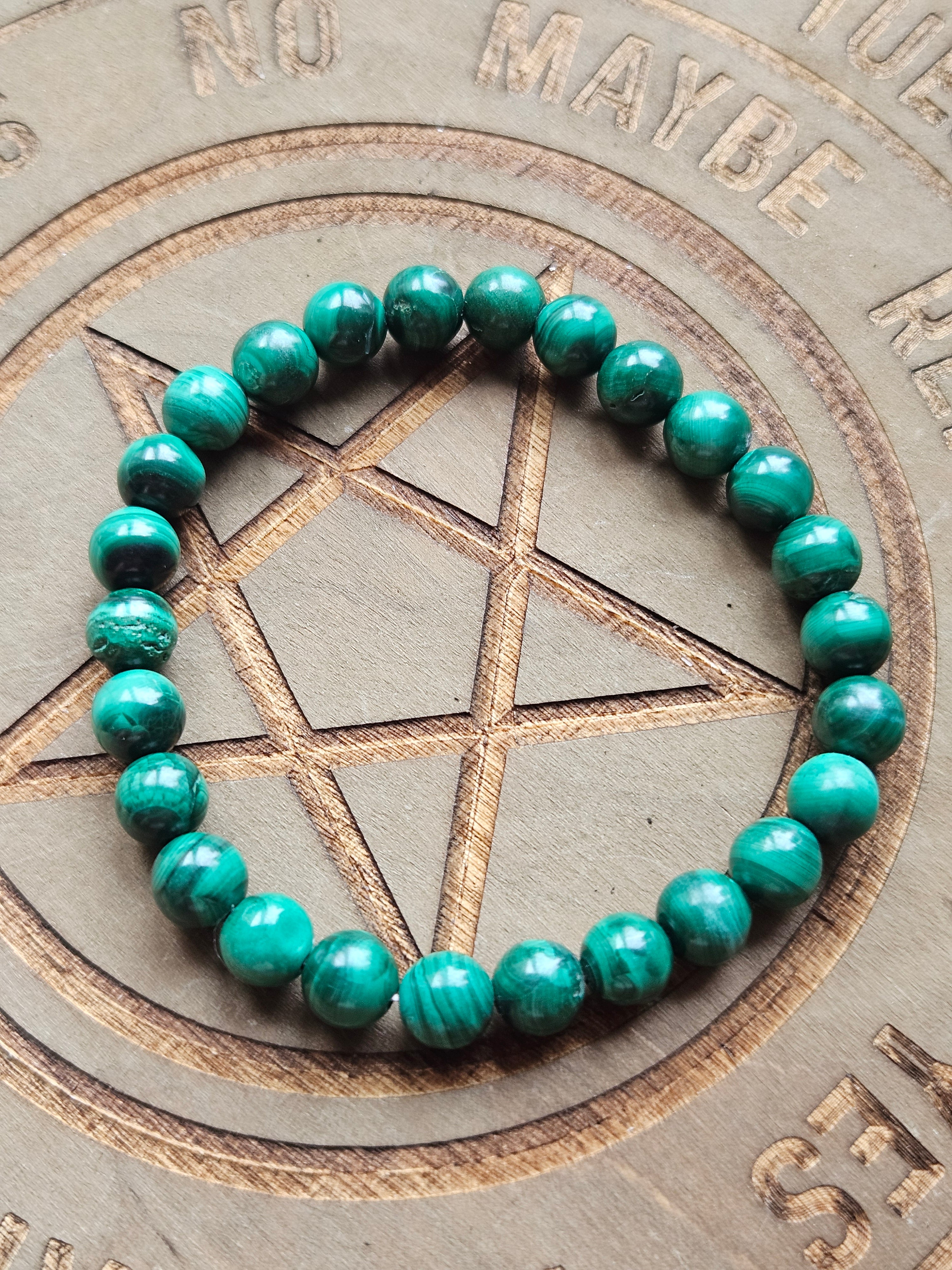 A beaded stretch bracelet made of polished green malachite beads, arranged in a circle on a wooden pentagram and altar board.