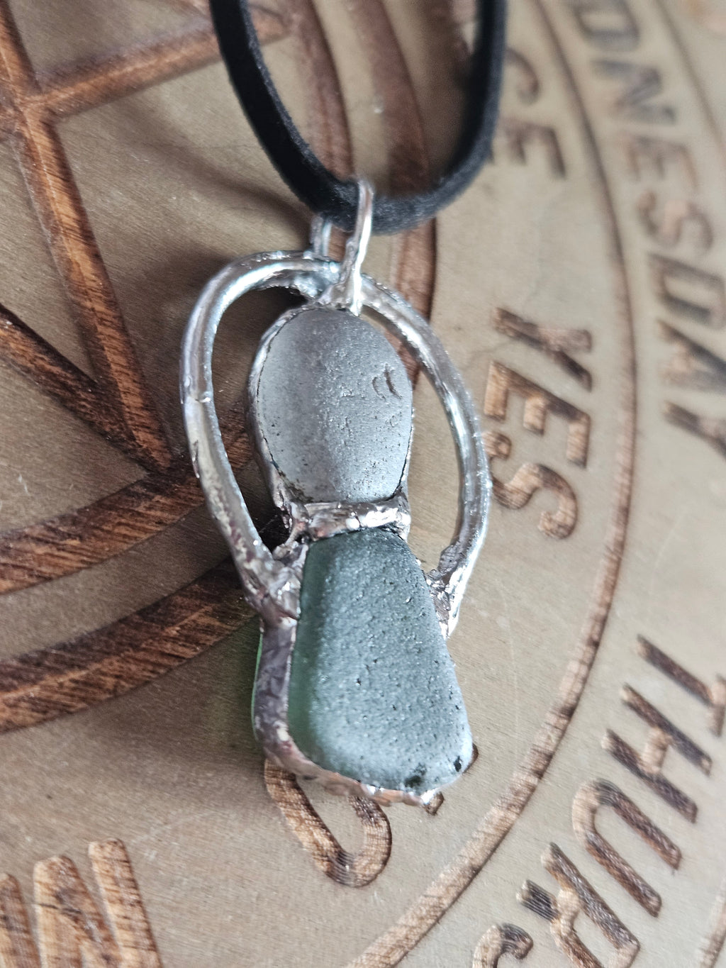 A close-up of a handmade pendant featuring a textured silver metal frame with two sea glass stones, one matte gray and one frosted light green, on a black cord.