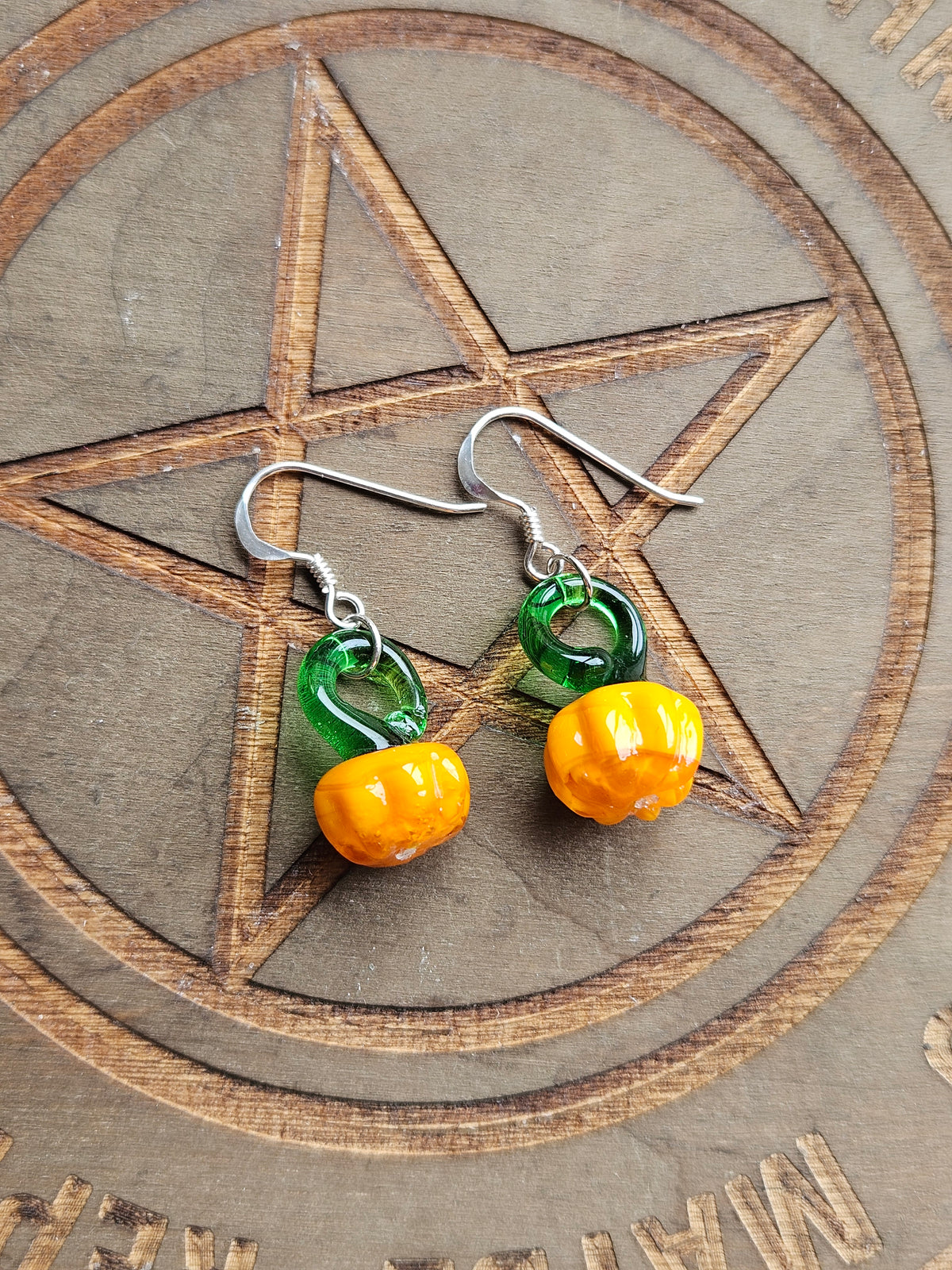 A pair of handmade dangle earrings featuring translucent green and orange glass beads shaped like small pumpkins, suspended from silver fishhook wires, displayed on a wooden pentagram board.
