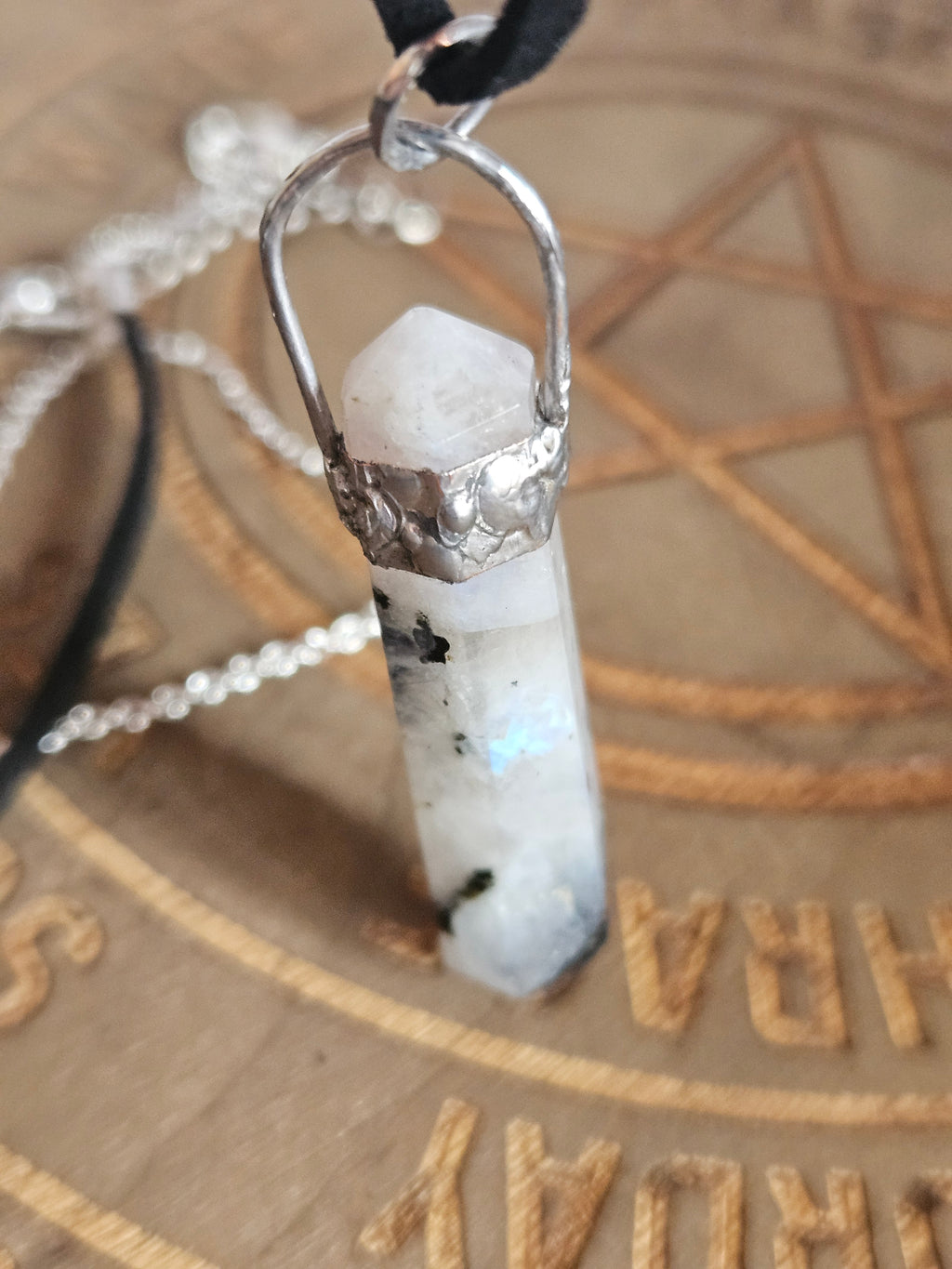 A close-up of a crystal pendant necklace featuring a rainbow moonstone point wrapped in textured silver metal, hanging from a silver chain over a wooden engraved background.