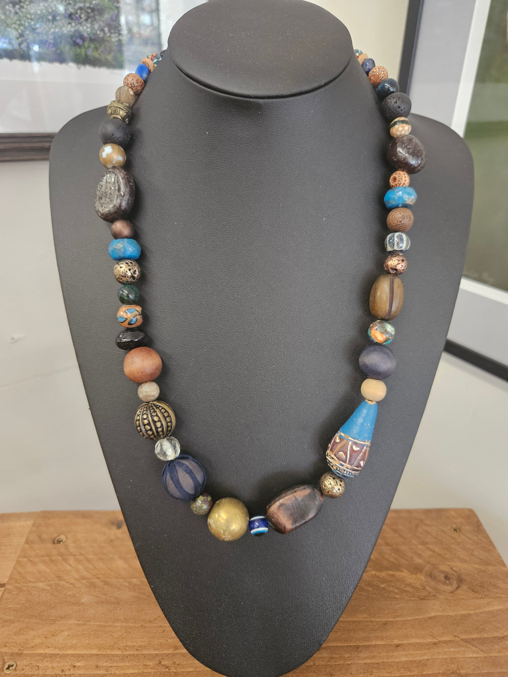 A multi-colored beaded necklace displayed on a matte black jewelry bust. The necklace features a variety of beads in blue, brown, gold, and gray, including patterned and textured glass beads, with a prominent blue teardrop-shaped bead near the bottom right.