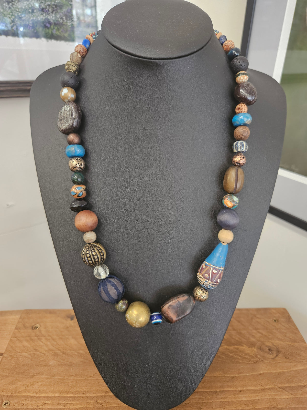 A multi-colored beaded necklace displayed on a matte black jewelry bust. The necklace features a variety of beads in blue, brown, gold, and gray, including patterned and textured glass beads, with a prominent blue teardrop-shaped bead near the bottom right.