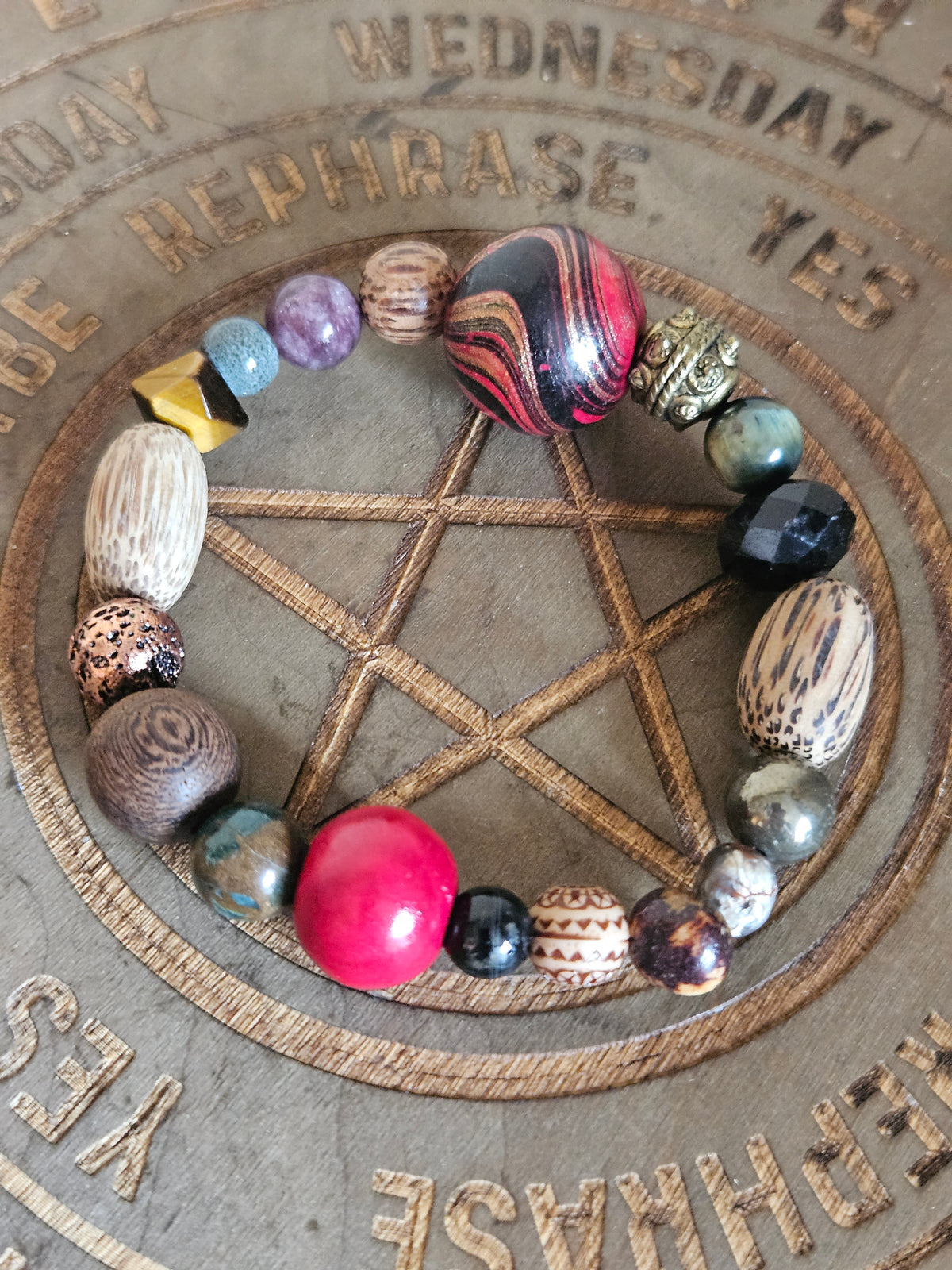 A handmade beaded boho bracelet arranged in a circle on a wooden pentacle board. The bracelet features a variety of beads including a prominent pink bead, striped red and black beads, patterned wood beads, and faceted stones in various colors, set against a rustic wooden background with engraved text.