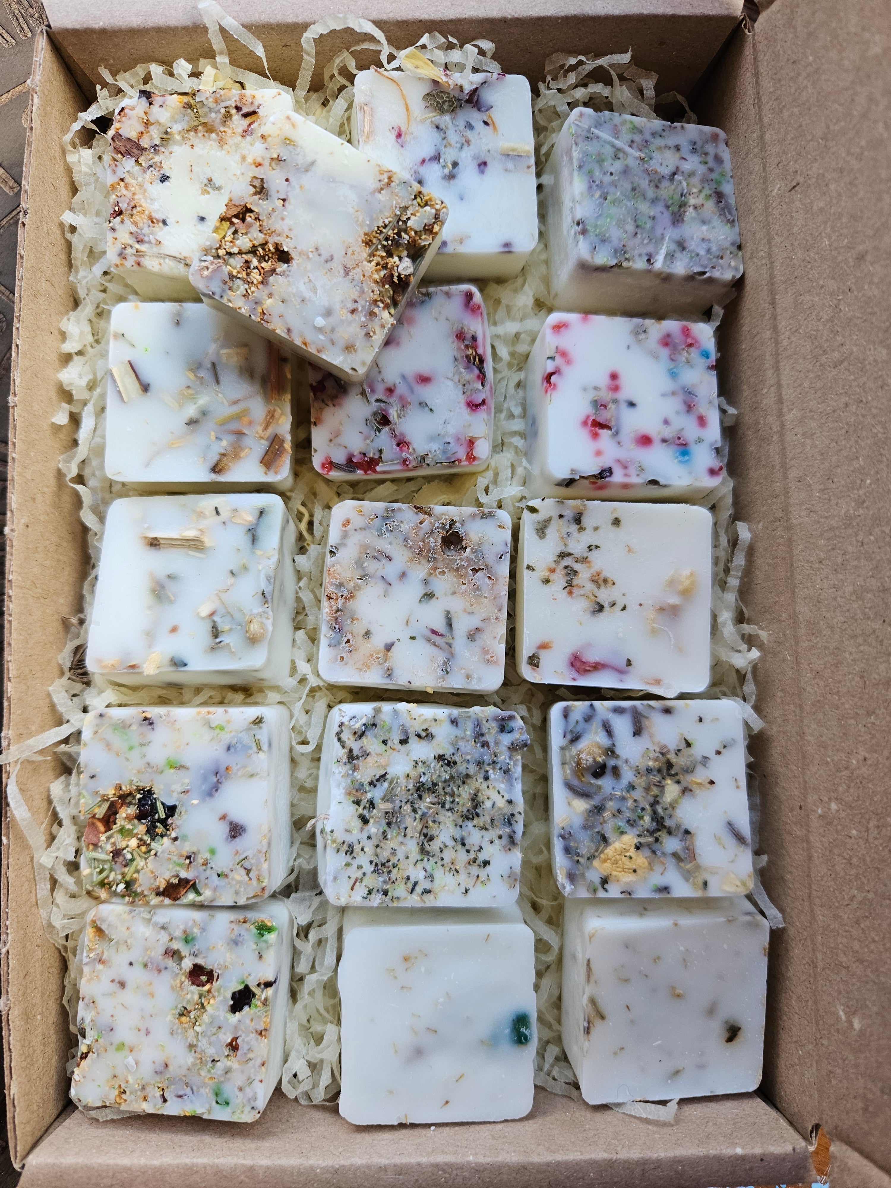 Several square handmade wax melts in off-white and cream tones, embedded with dried botanicals, seeds, and herbs, arranged in shredded paper inside a cardboard box.