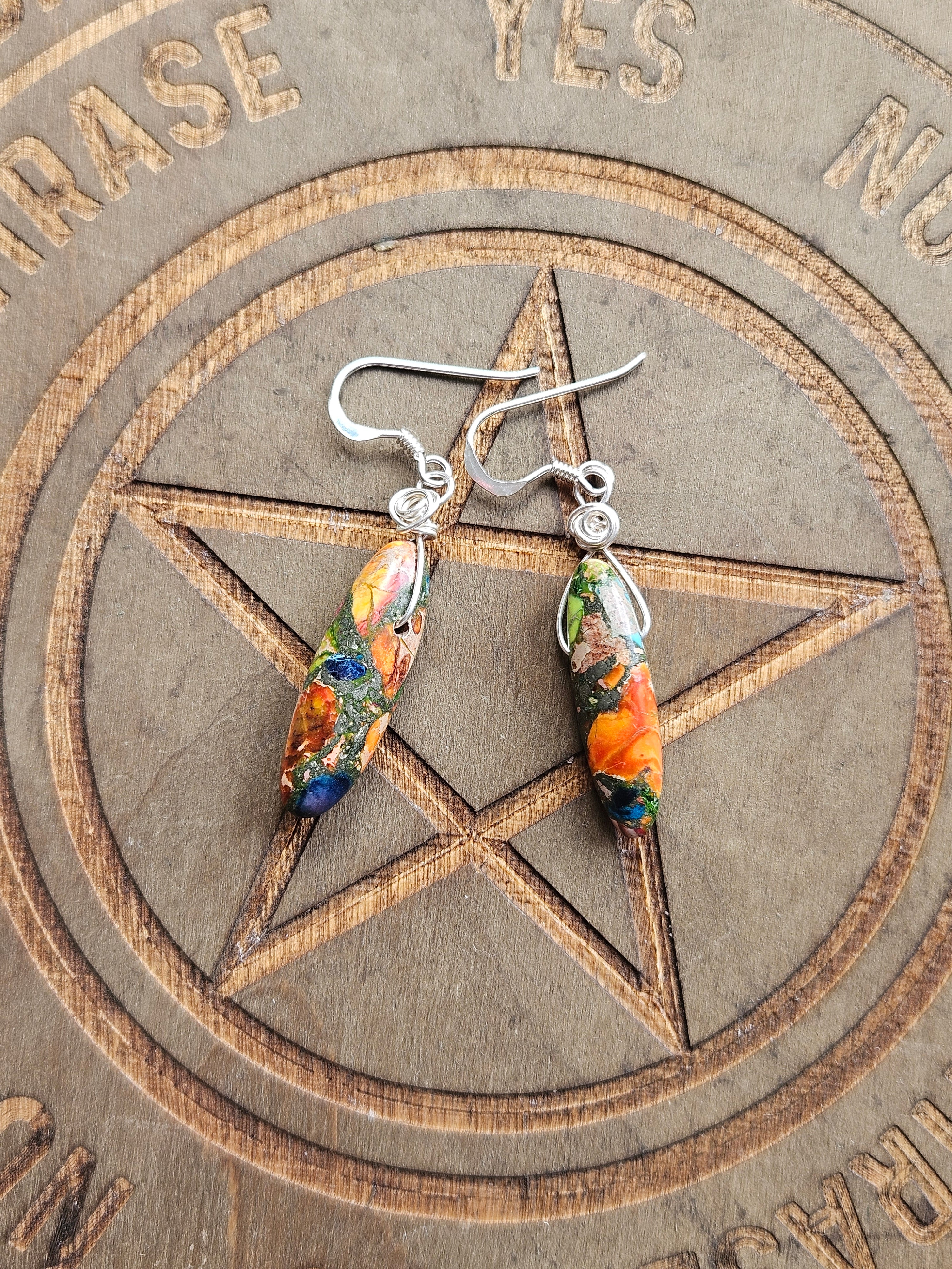 A pair of handmade dangle earrings with silver fishhook wires and colorful marbled Sediment Jasper beads in orange, green, blue, and yellow, displayed on a carved wooden pentagram board.