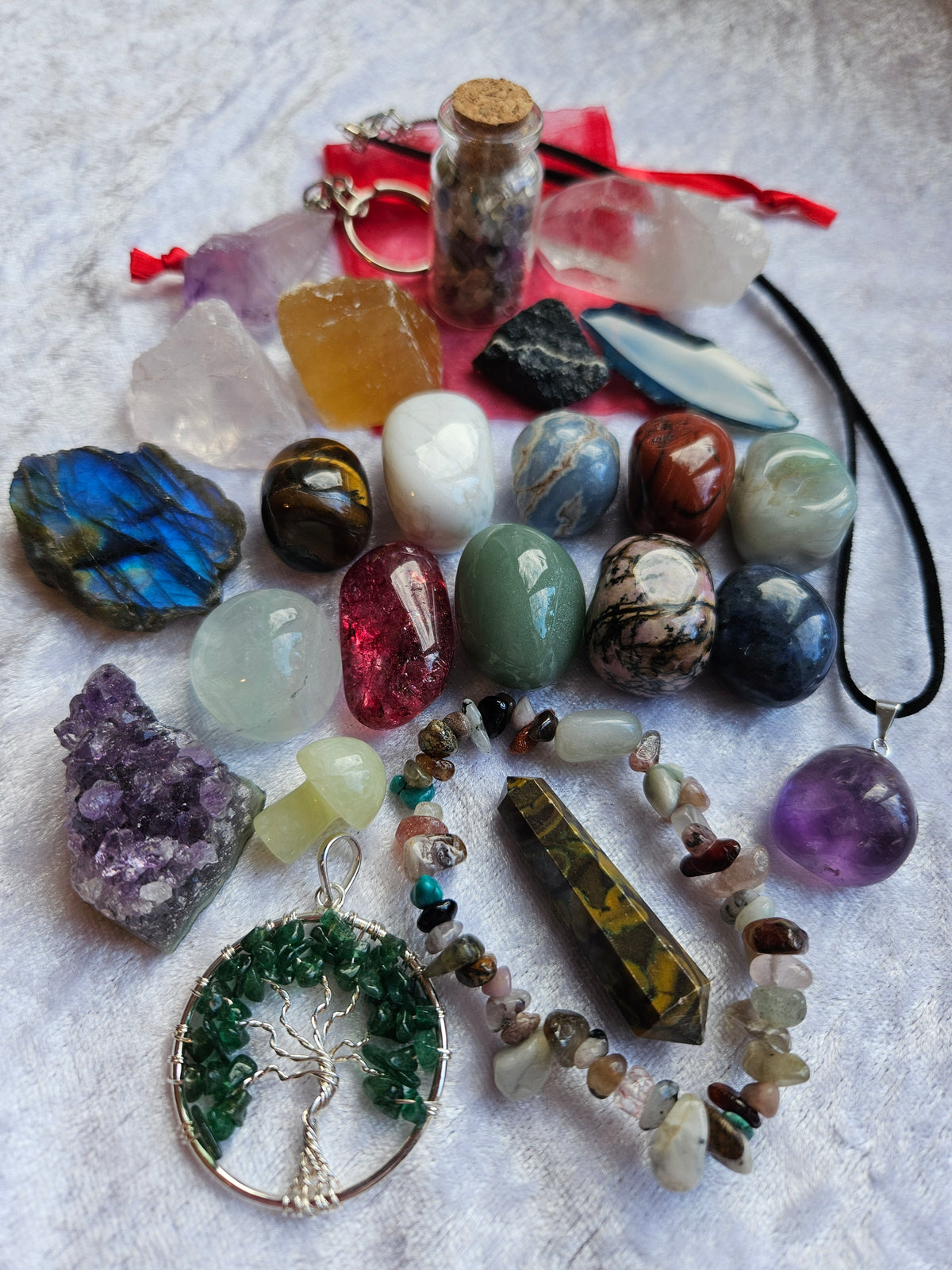 Advent calendar contents, a collection of colorful raw gemstones, polished stones, crystals, and jewelry pieces arranged on a white textured fabric background. The assortment includes a tree of life pendant, a purple crystal cluster, a green crystal pendant, a crystal chip bracelet, and various other raw stones and a small vial with a cork stopper.