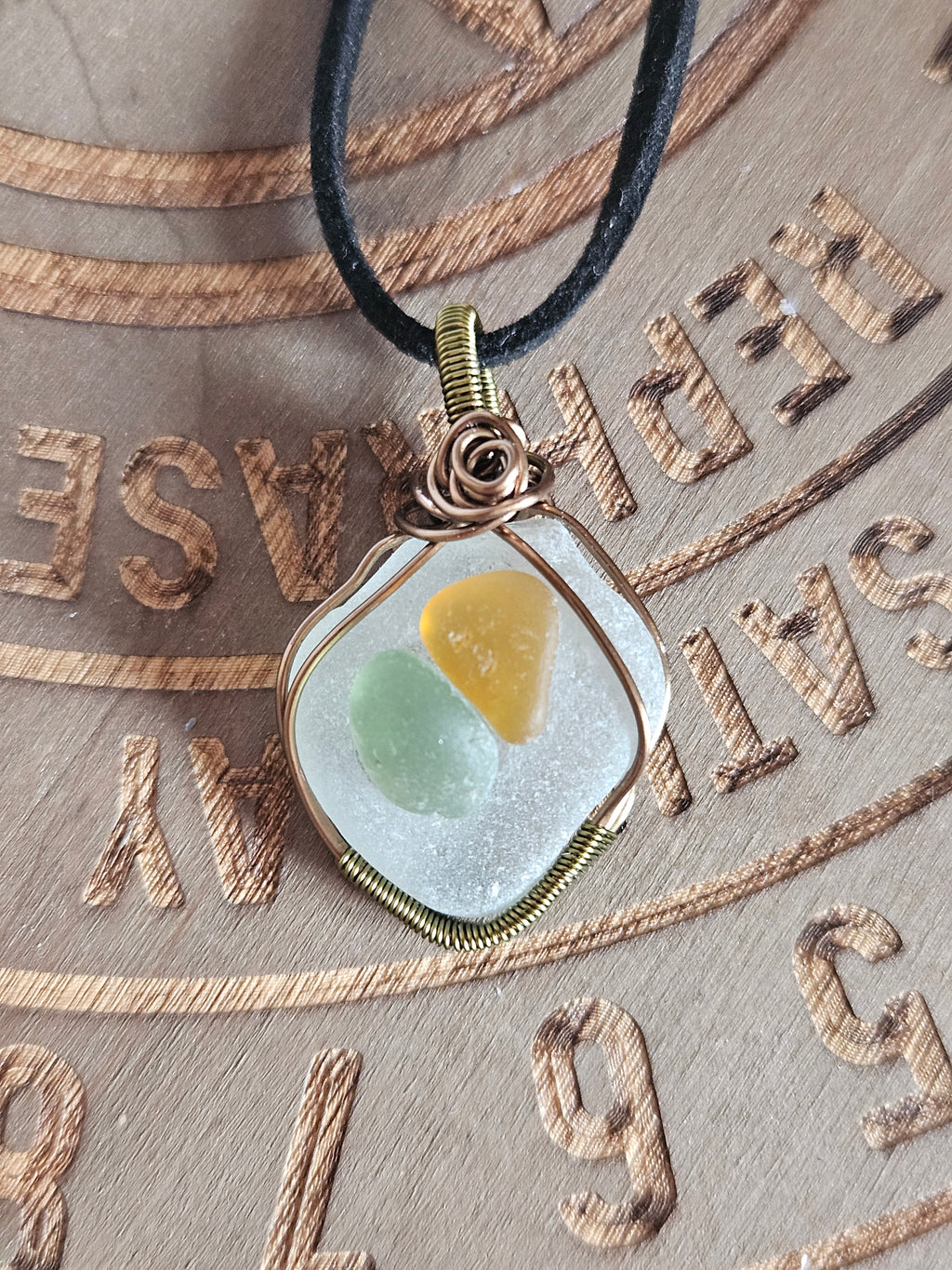 A sea glass pendant necklace with a black cord, featuring a white frosted sea glass cabochon containing yellow and green inclusions, wrapped in a rustic copper wire setting on a textured background.