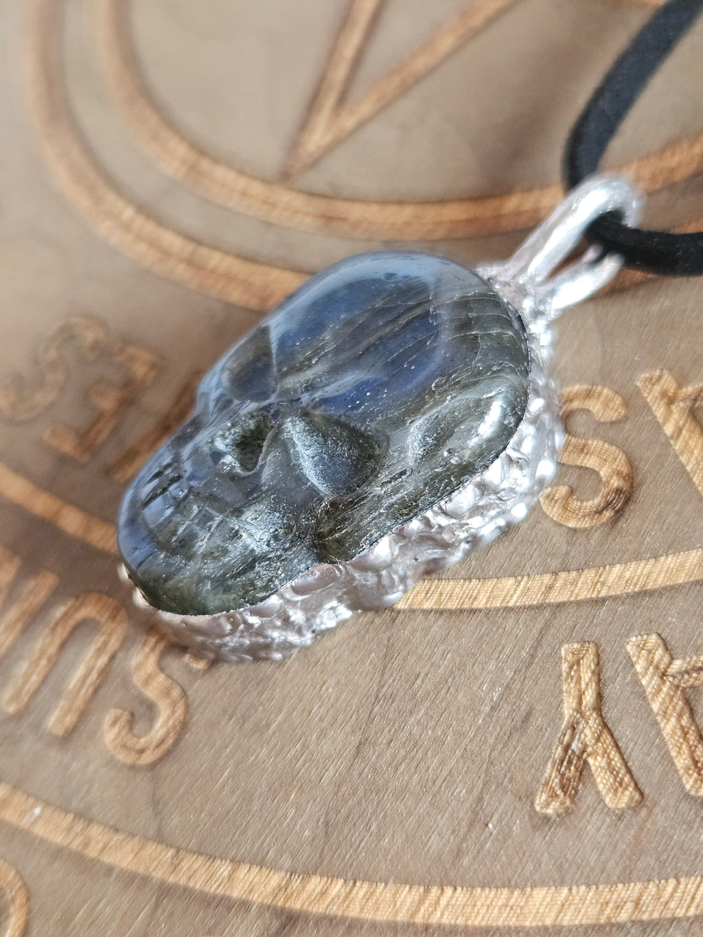 A close-up of a carved skull pendant made from a dark gray to black stone, set in a textured silver-tone metal frame and hanging from a black cord, resting on a wooden background with engraved symbols.