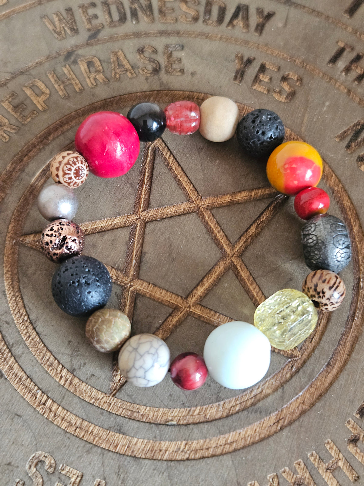 A beaded boho bracelet arranged in a circle on a wooden surface featuring an engraved pentagram and mystical text. The bracelet contains a variety of beads in pink, red, black, white, yellow, brown, and metallic gold.