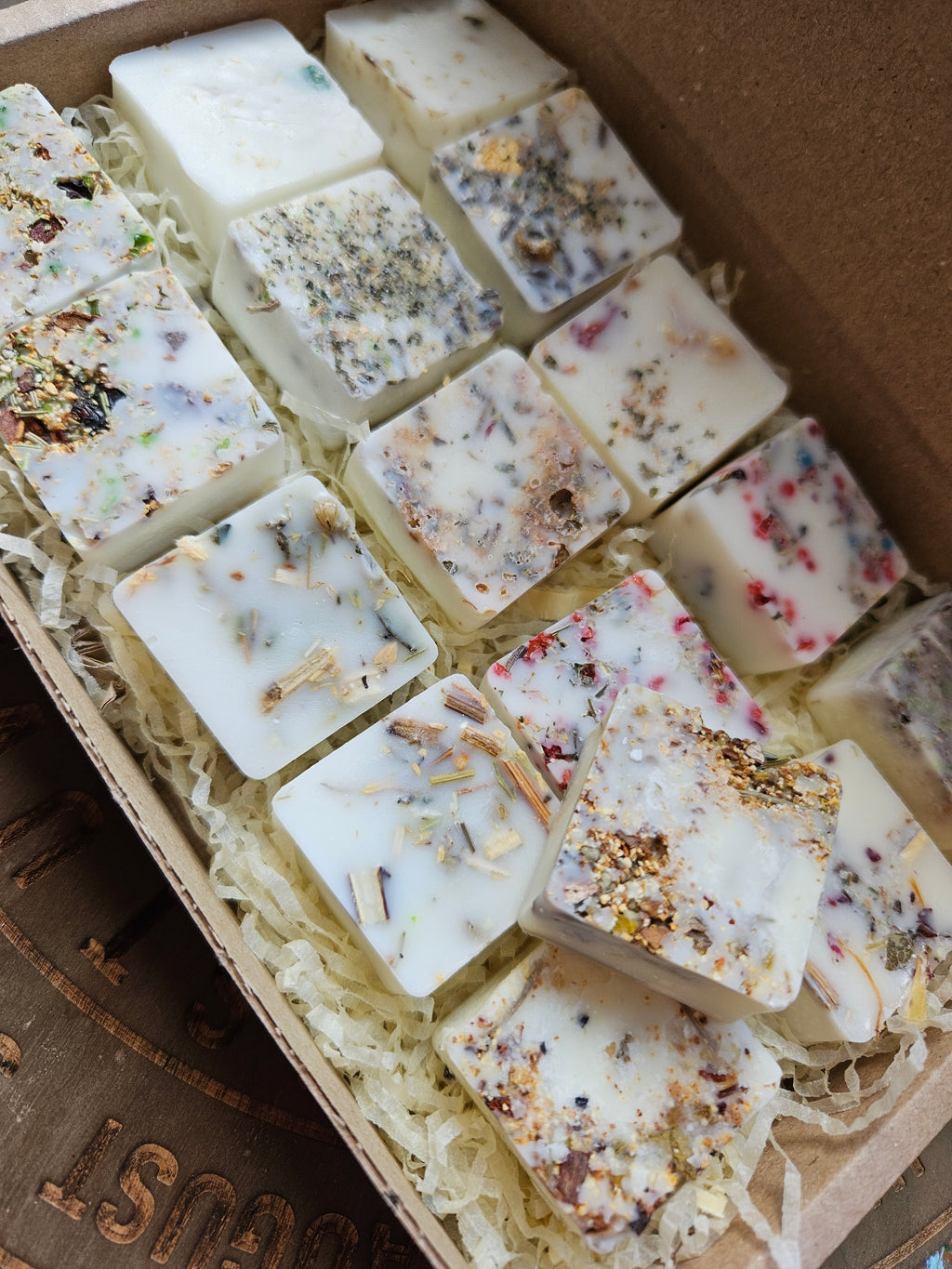 Several square handmade wax melts in off-white and cream tones, embedded with dried botanicals, seeds, and herbs, arranged in shredded paper inside a cardboard box.