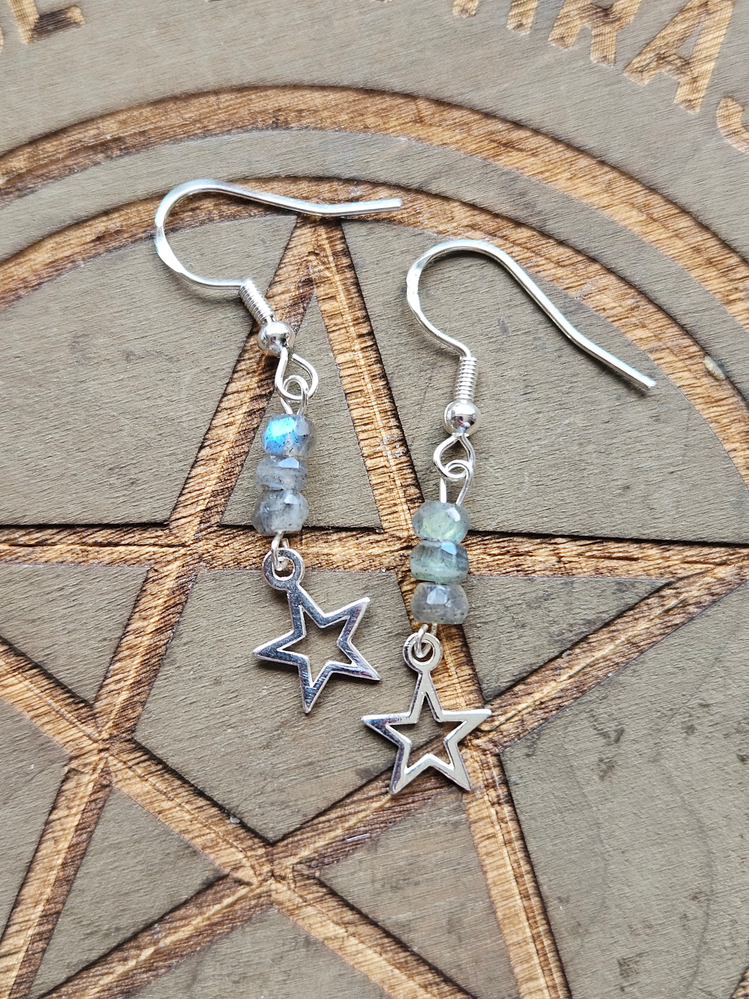 A pair of sterling silver dangle earrings with star charms and small iridescent Labradorite beads, displayed on a wooden surface with an engraved pentagram.