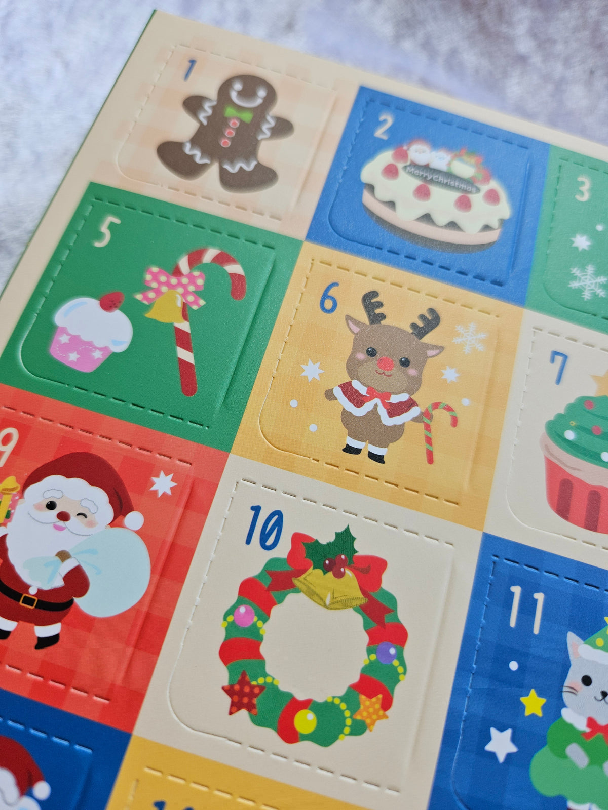A close-up of a colorful Christmas advent calendar page showing numbered square doors with festive illustrations like a wreath, Santa, gingerbread, candy canes, and desserts.
