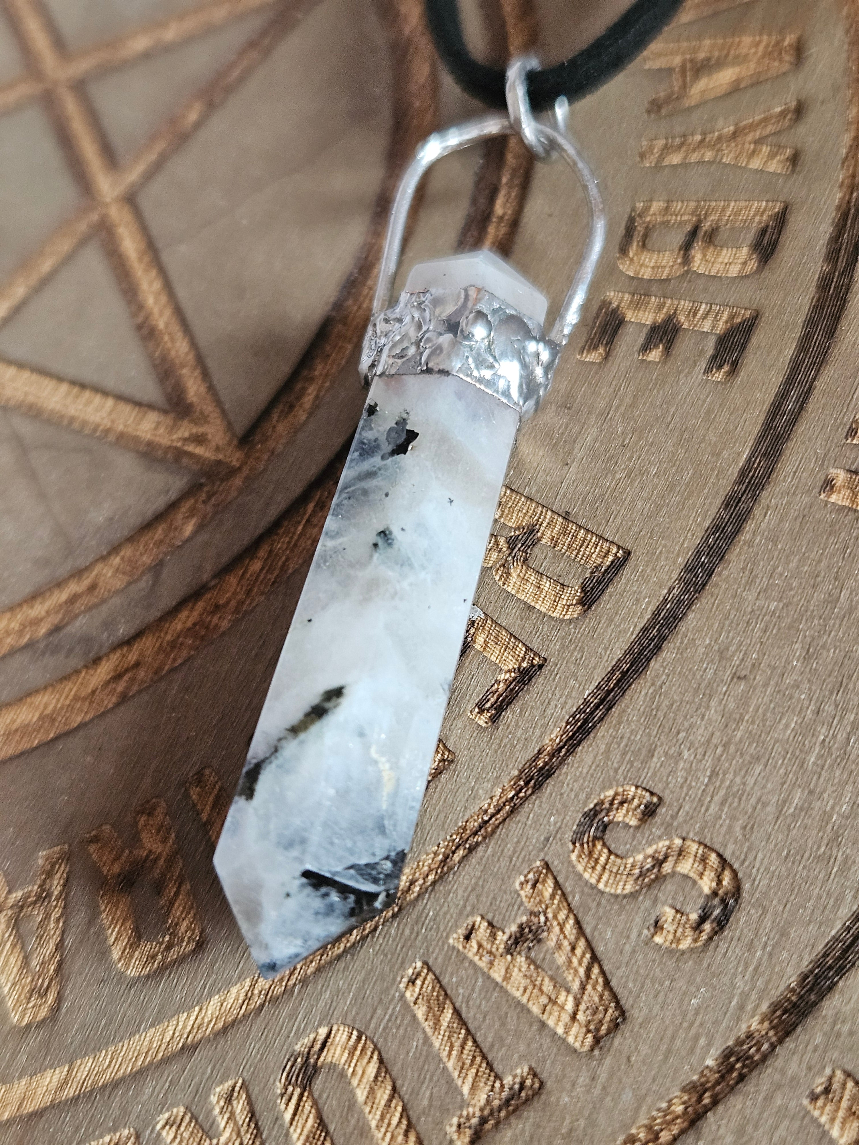 A polished moonstone crystal pendant with black inclusions, suspended from a silver-colored metal loop on a black cord, resting on a wooden engraved background.