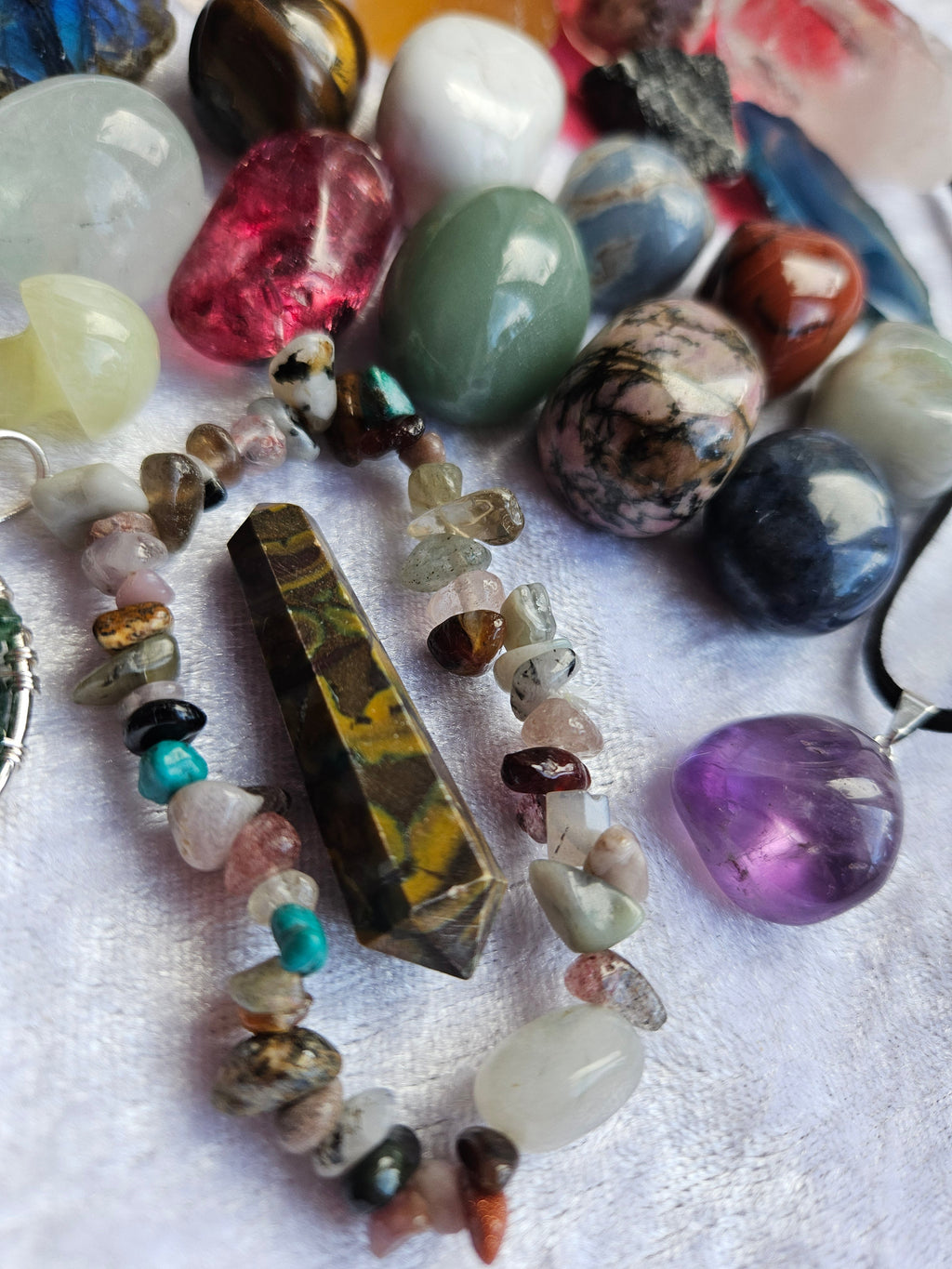 A beaded bracelet made of polished gemstone beads and a pointed crystal, displayed among other colorful gemstones on a white textured fabric background.