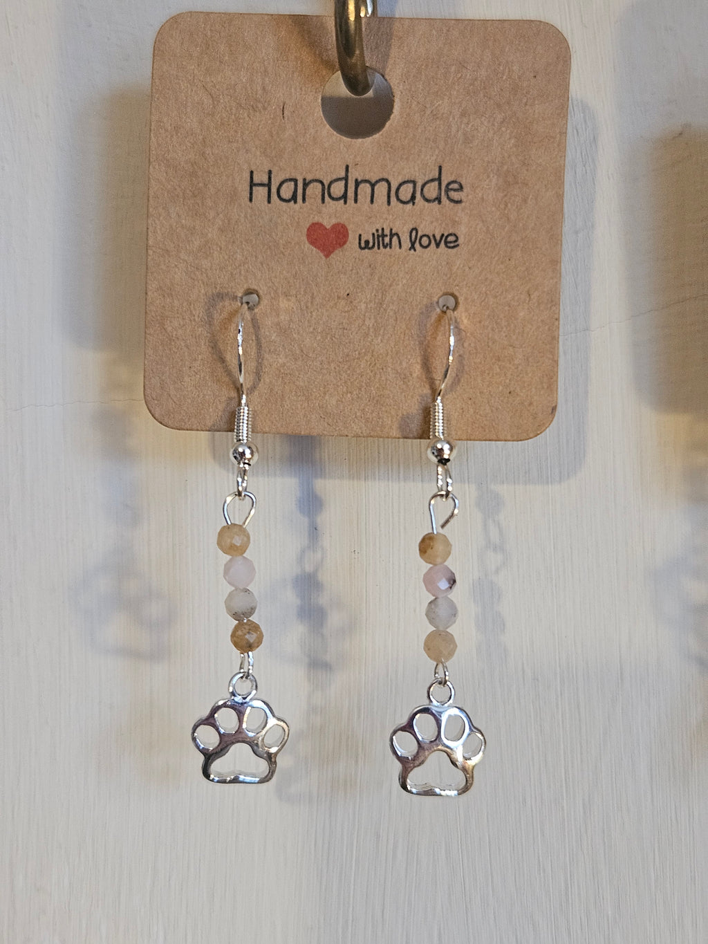Pair of paw-shaped earrings hanging from a 'Handmade with Love' tag on a wooden surface.