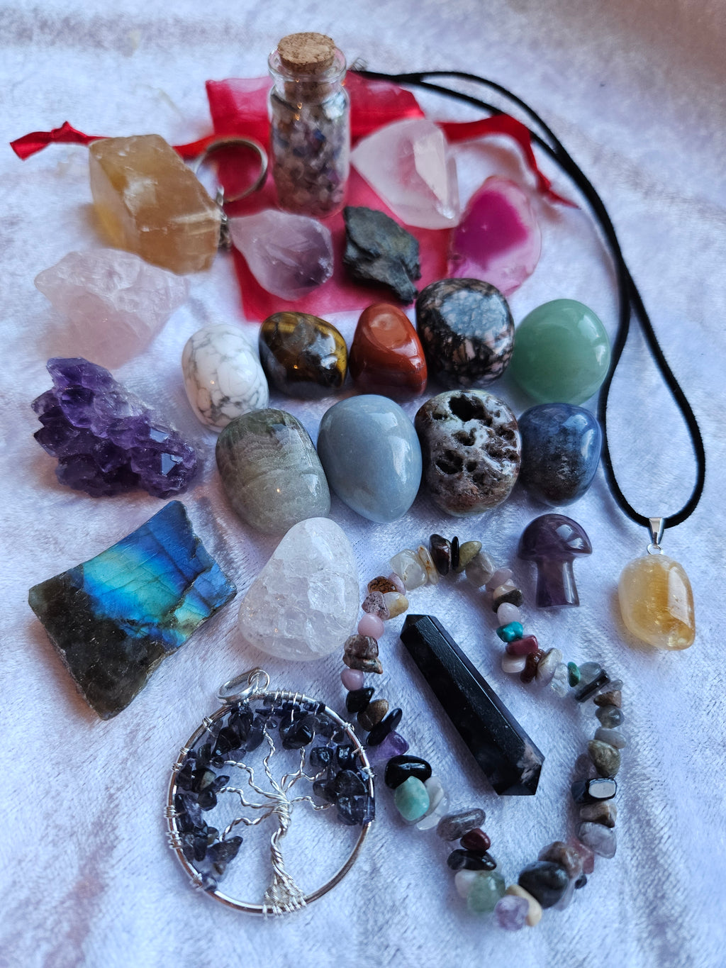 Advent calendar contents, a collection of colorful raw gemstones, polished stones, crystals, and jewelry pieces arranged on a white textured fabric background. The assortment includes a tree of life pendant, a purple crystal cluster, a green crystal pendant, a crystal chip bracelet, and various other raw stones and a small vial with a cork stopper.