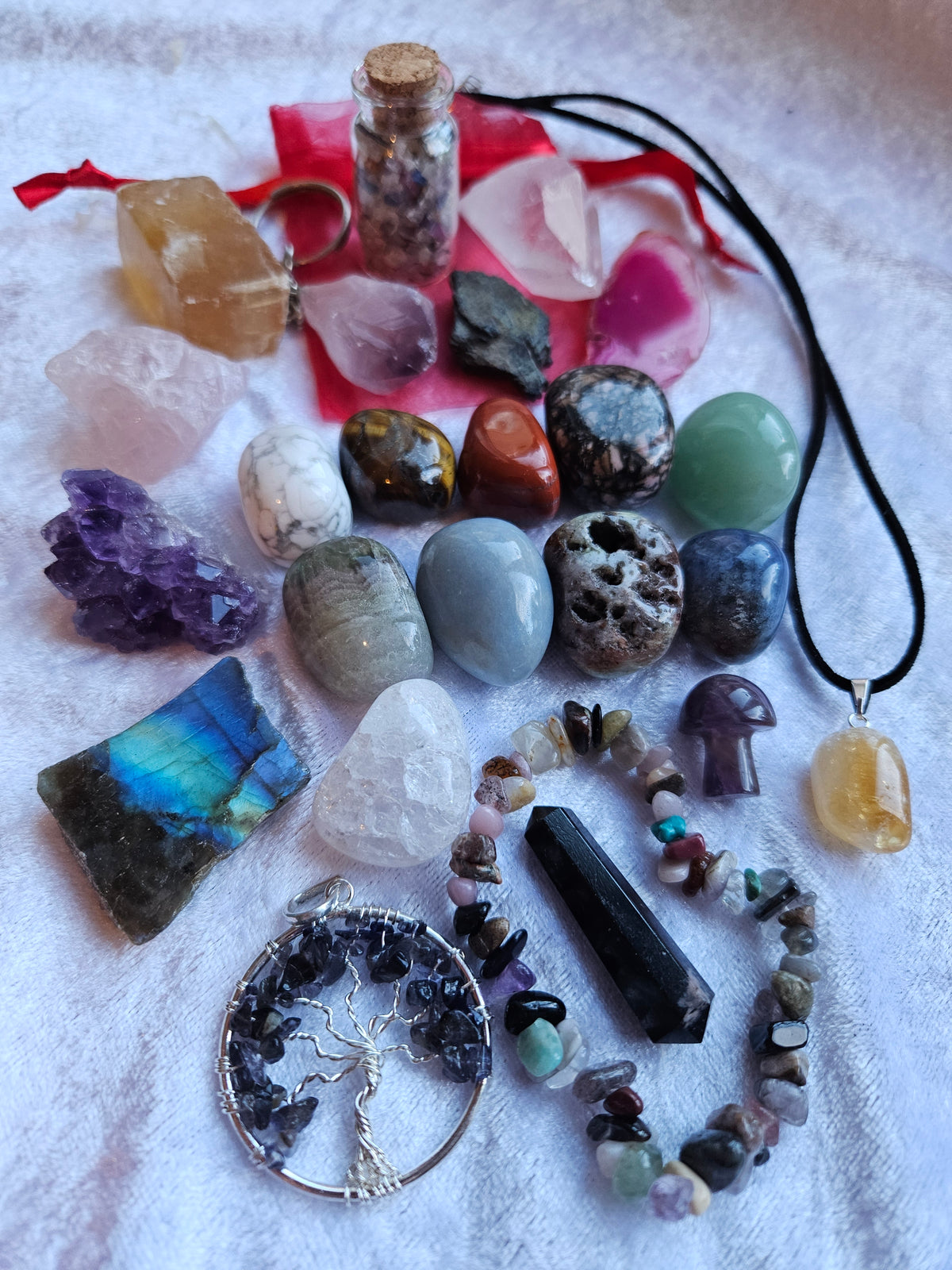 Advent calendar contents, a collection of colorful raw gemstones, polished stones, crystals, and jewelry pieces arranged on a white textured fabric background. The assortment includes a tree of life pendant, a purple crystal cluster, a green crystal pendant, a crystal chip bracelet, and various other raw stones and a small vial with a cork stopper.