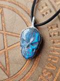 A skull-shaped pendant with a metallic blue and silver finish, hanging from a black cord, set against a wooden background with engraved text and a pentagram design.