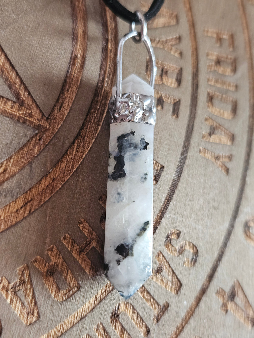 A white and black speckled crystal point pendant with a silver metal cap and black loop, resting on a brown engraved wooden surface.