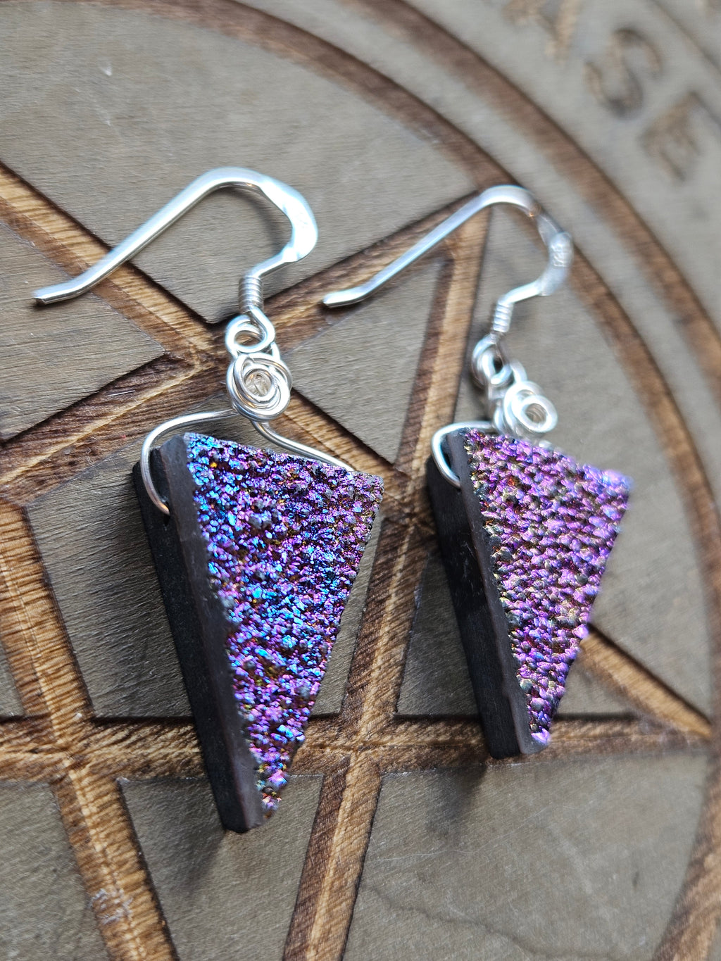 A pair of handmade  triangular dangle earrings with iridescent purple Drusy Quartz pendants and sterling silver fishhook wires, displayed on a wooden pentagram board.