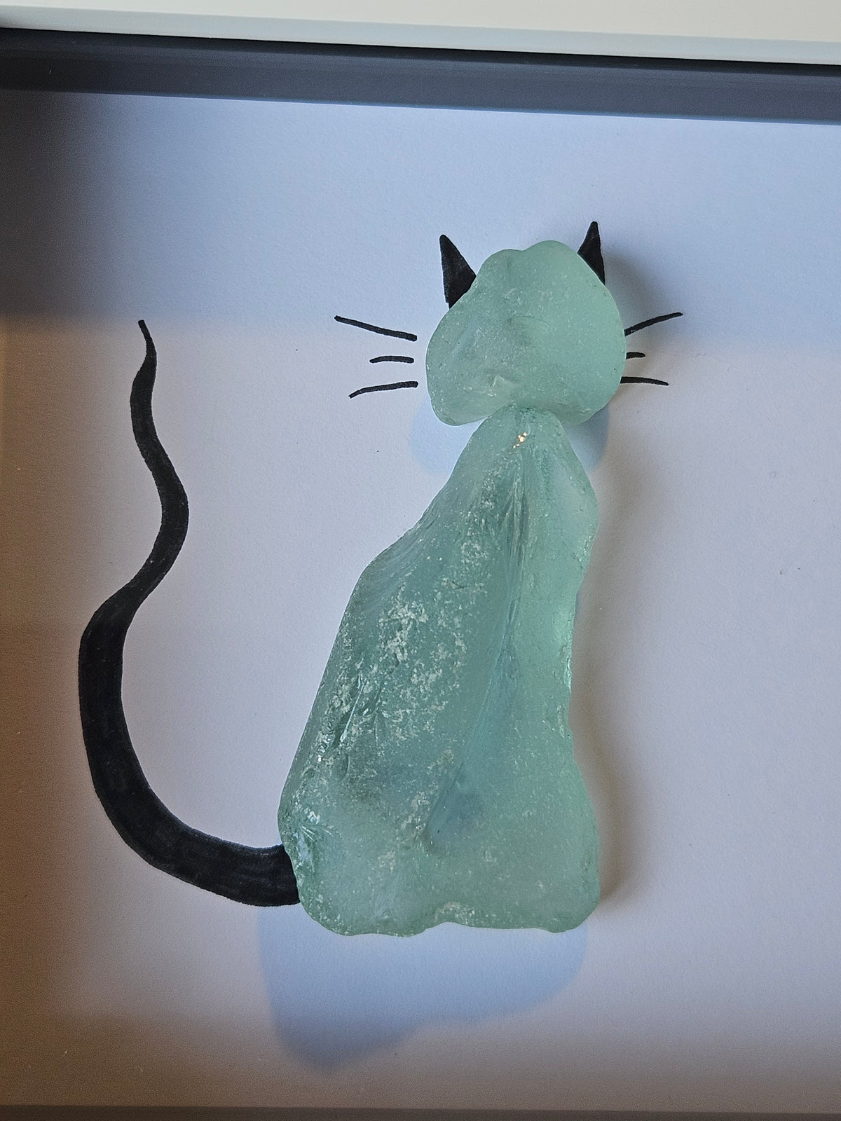 A sea glass cat figurine crafted from textured light green glass, featuring black hand-painted whiskers, ears, and a long tail, displayed inside a clear glass frame.